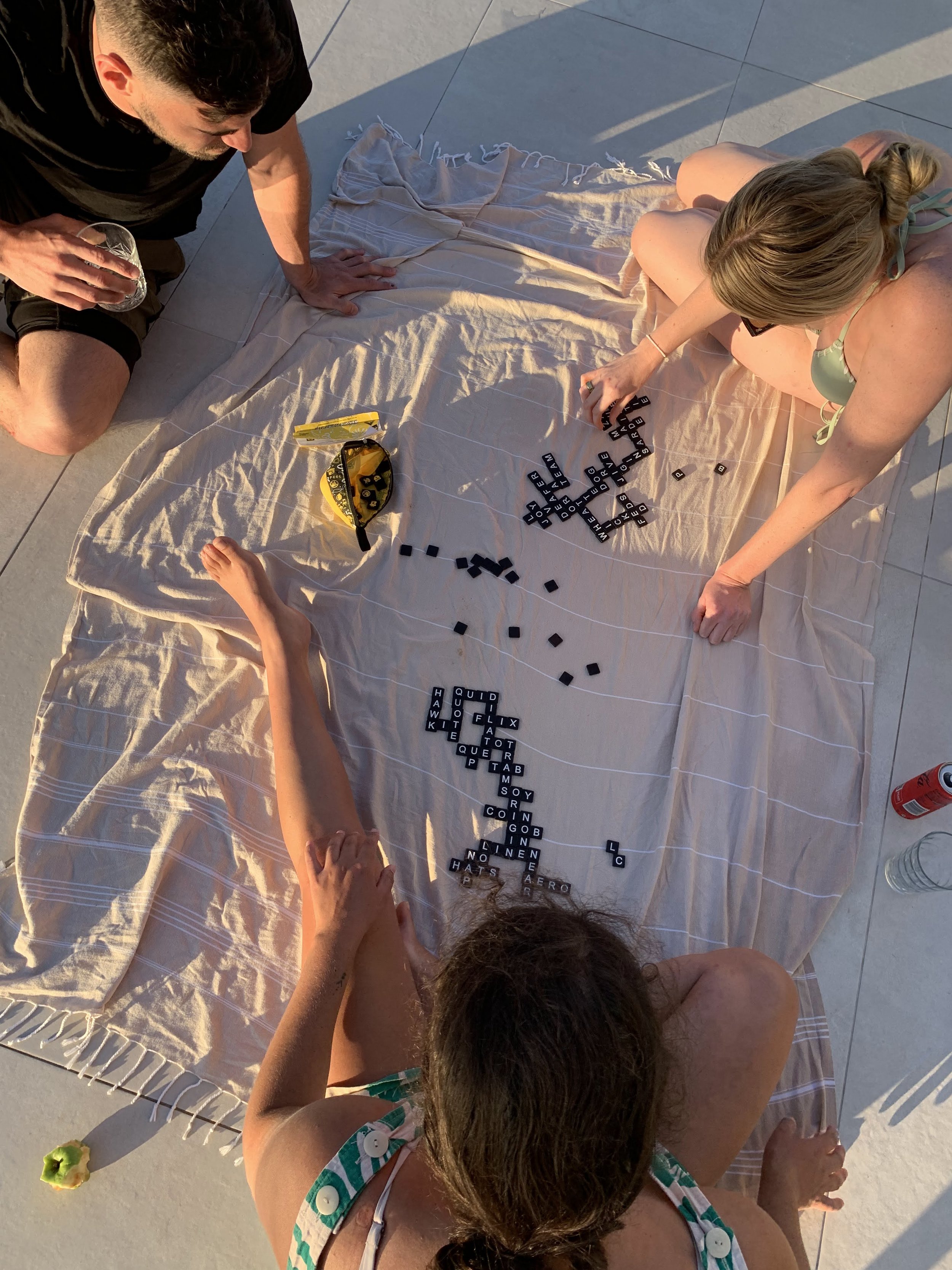 Three people sitting on a blanket outdoors playing a word game with black letter tiles. There's a yellow game pouch nearby, as well as drinks and a green apple core on the floor.