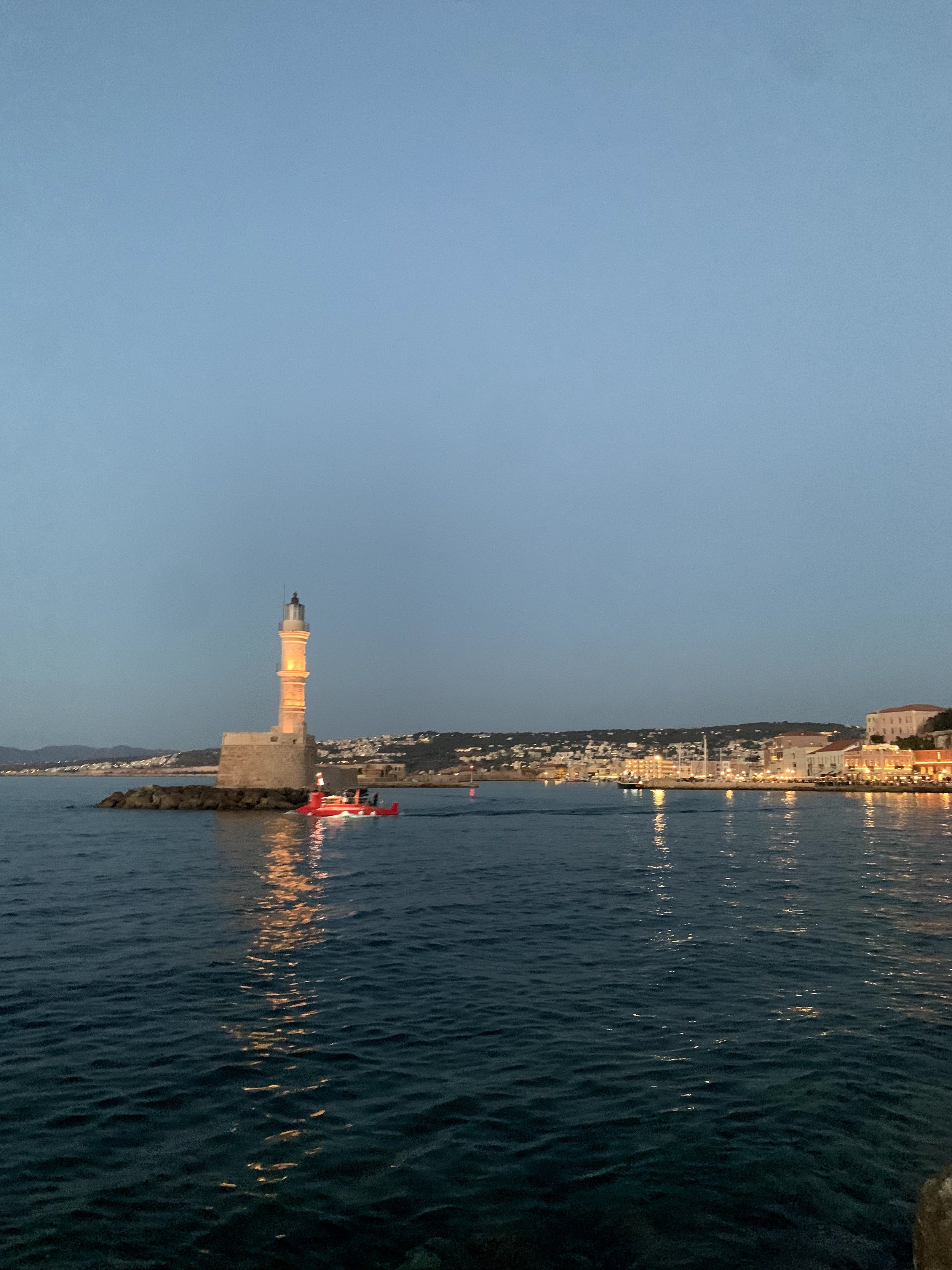 Lighthouse at dusk in Chania harbor with city lights reflecting on the water.