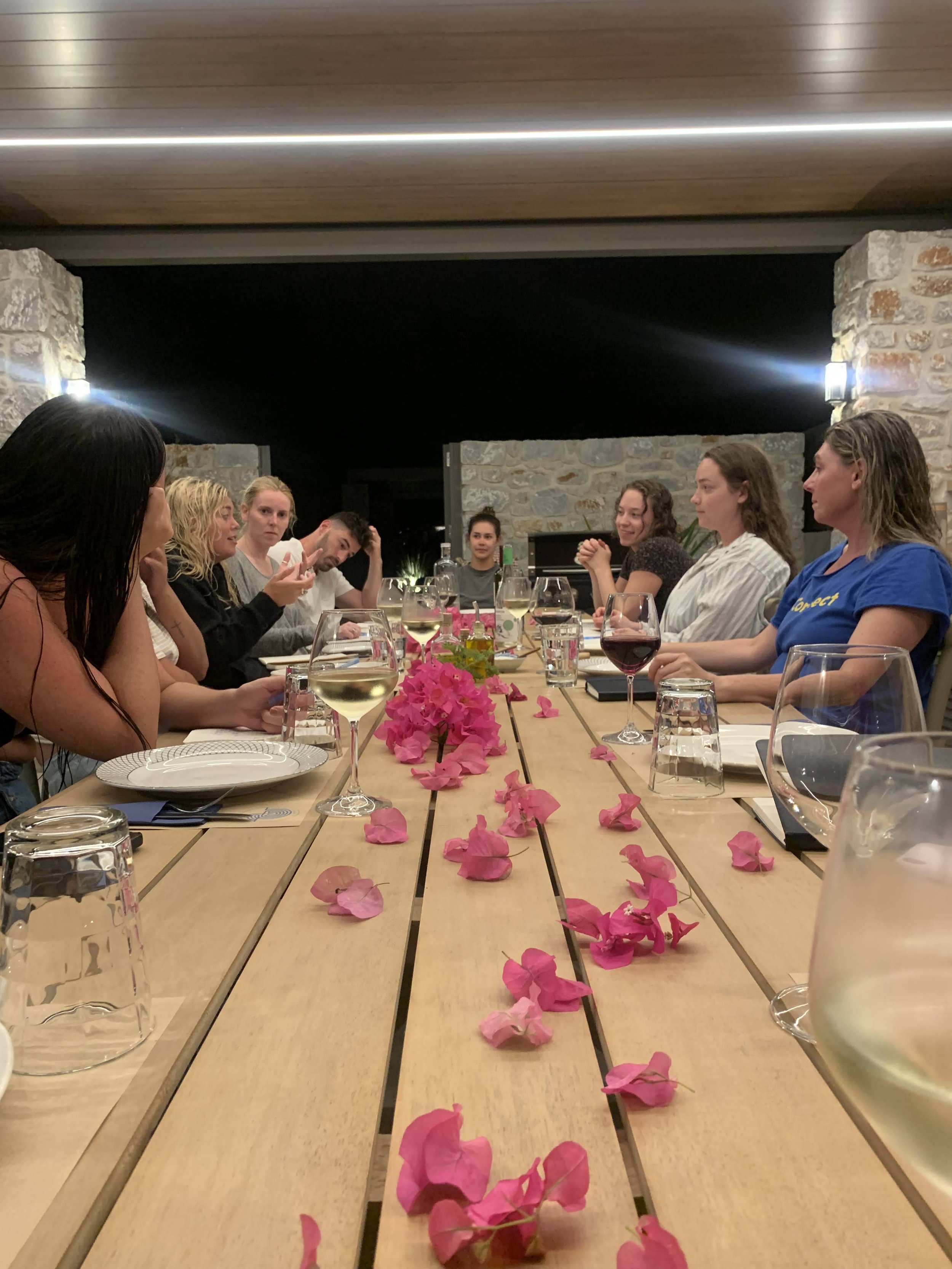Group of people sitting around a long wooden table decorated with pink flowers, sharing a meal in a stone-walled outdoor setting at night.