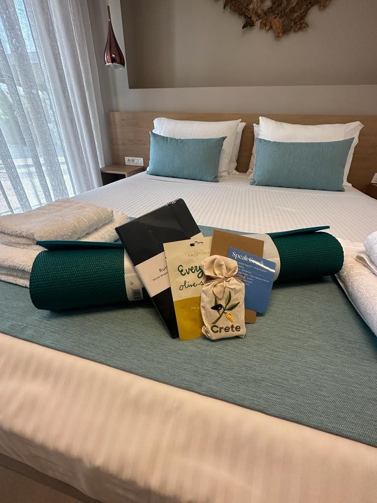Hotel room with a neatly made bed featuring two white pillows and teal cushion. On the bed, there are several items including a rolled green yoga mat, brochures, a small bag labeled "Crete," and a stacked white towel. A curtain and modern wall decor
