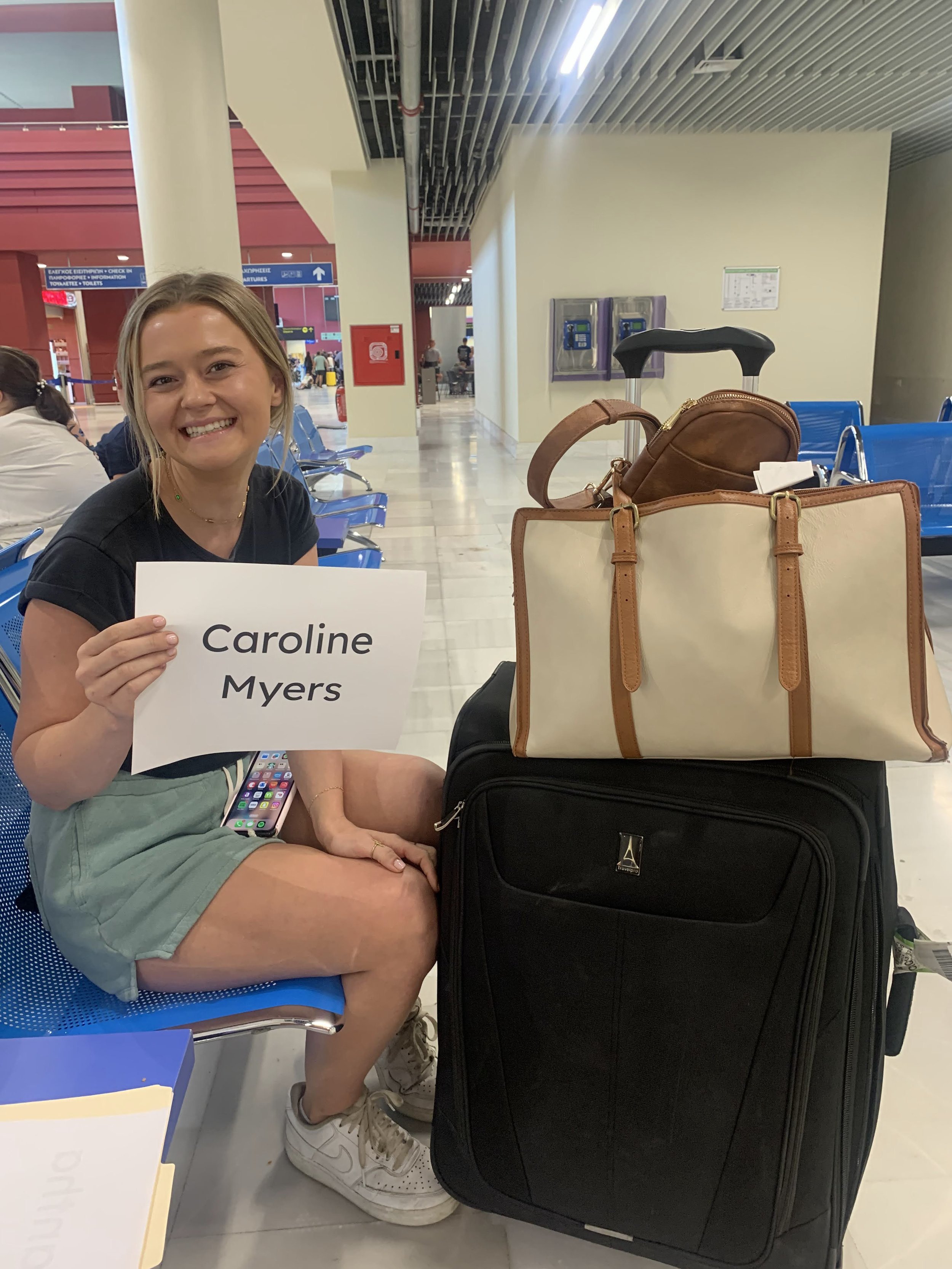 Person sitting in an airport holding a sign that reads 'Caroline Myers' next to a suitcase and a bag.
