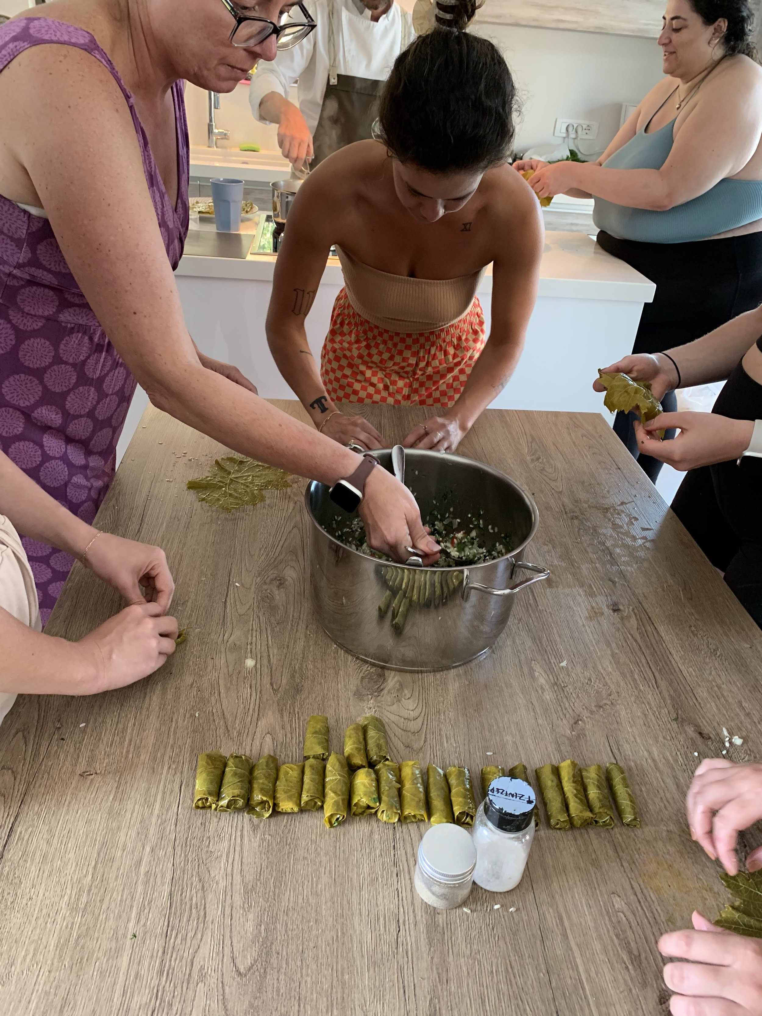 People preparing dolmas, rolling grape leaves with rice filling, around a table with spices and a pot.