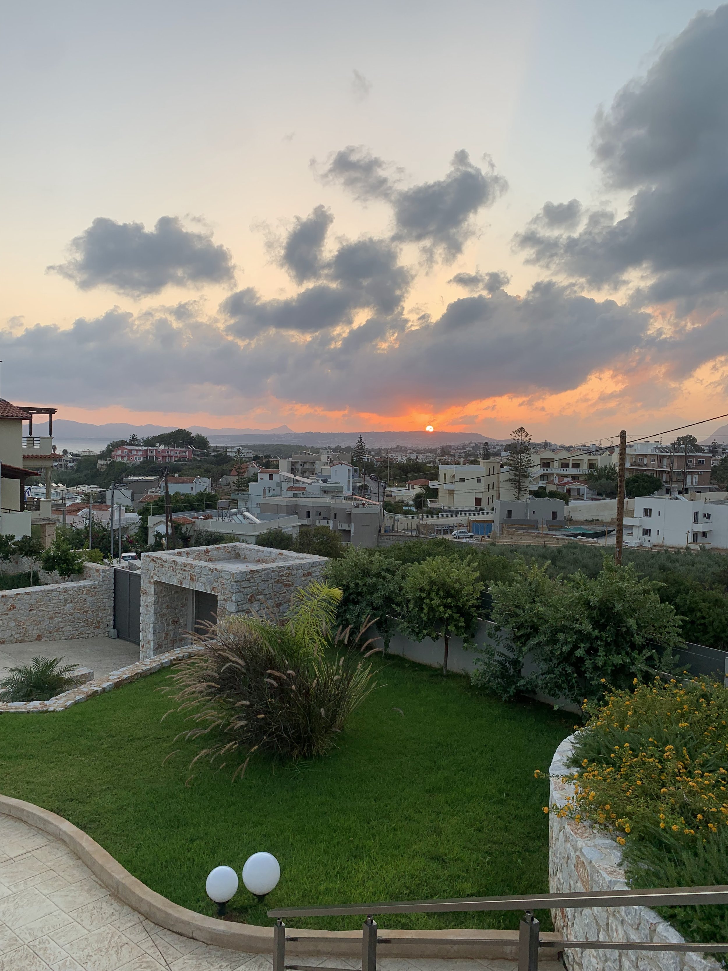 Sunset view over a residential neighborhood with green lawns and stone walls, under a sky with scattered clouds.