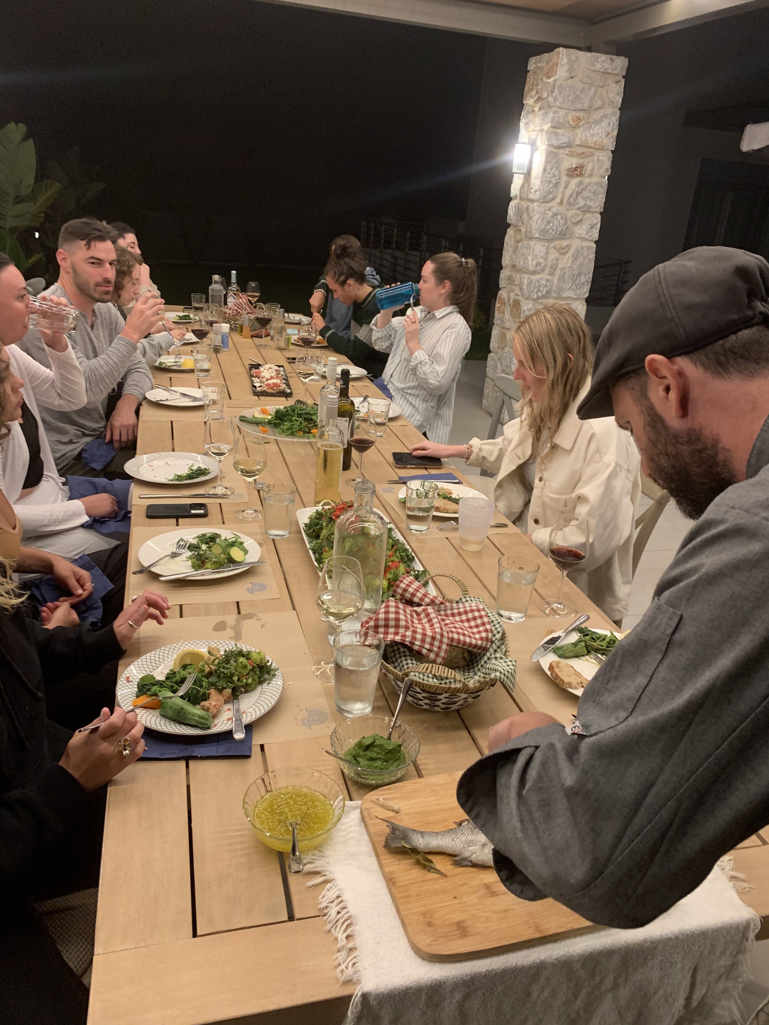 People gathered around a long wooden table enjoying a meal, with plates of food, wine glasses, and bread in baskets. A person is carving a fish on a cutting board.