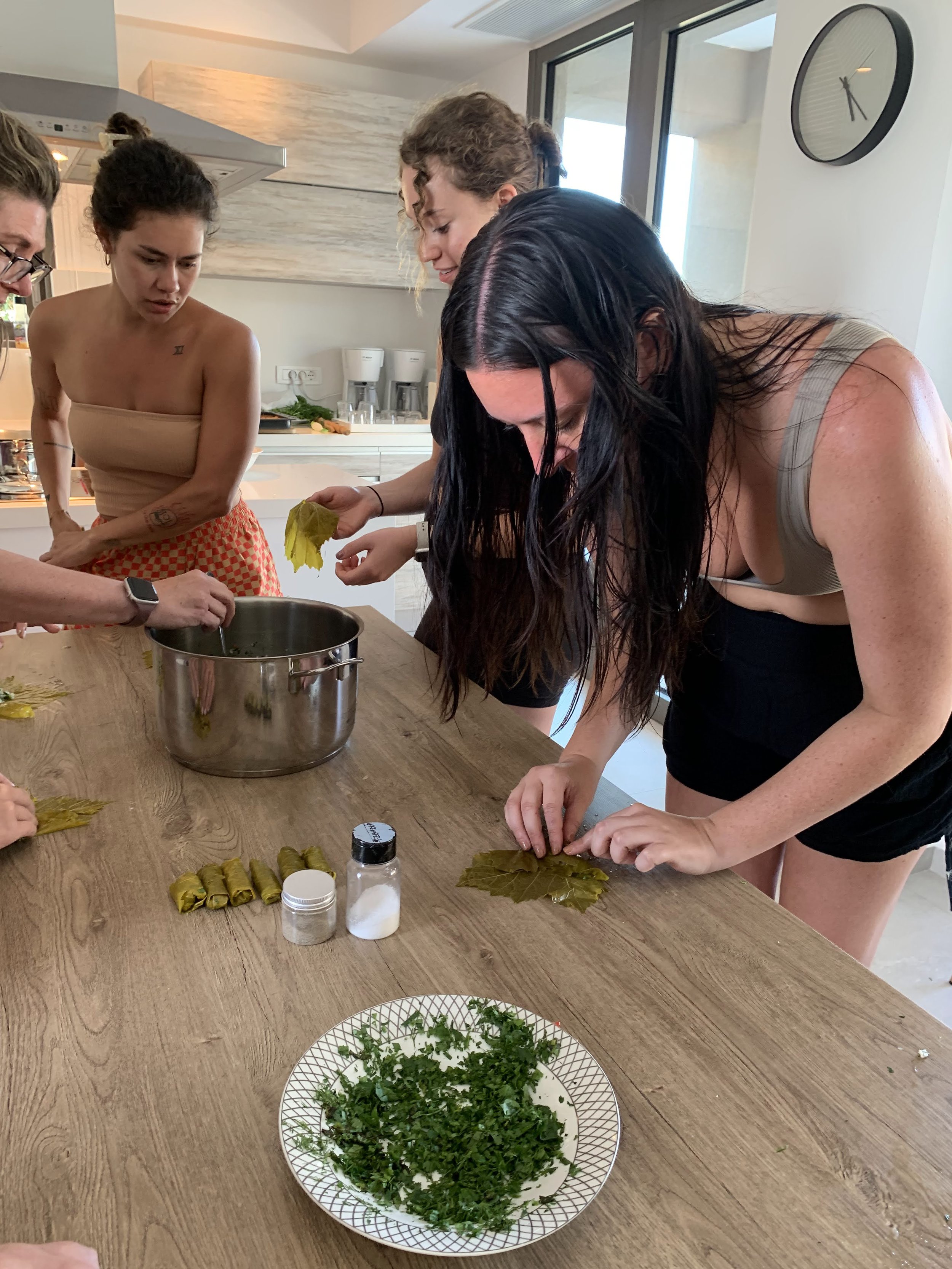 People rolling grape leaves at a kitchen table, with a pot and ingredients like salt and chopped herbs nearby.