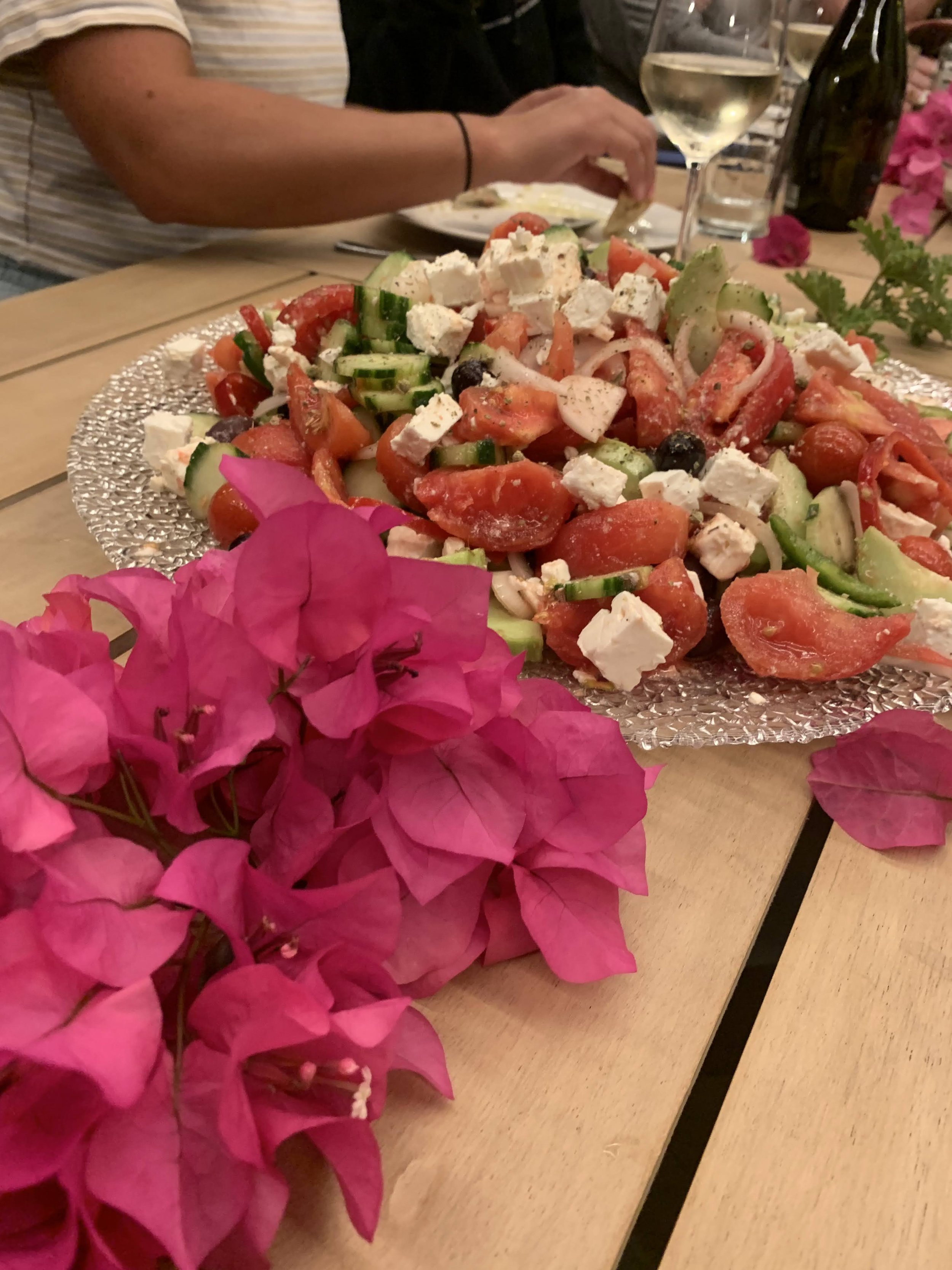 A Greek salad with tomatoes, cucumbers, feta cheese, olives, and herbs on a glass platter. It is placed on a wooden table alongside bright pink flowers, a glass of white wine, and two people not fully visible in the background.