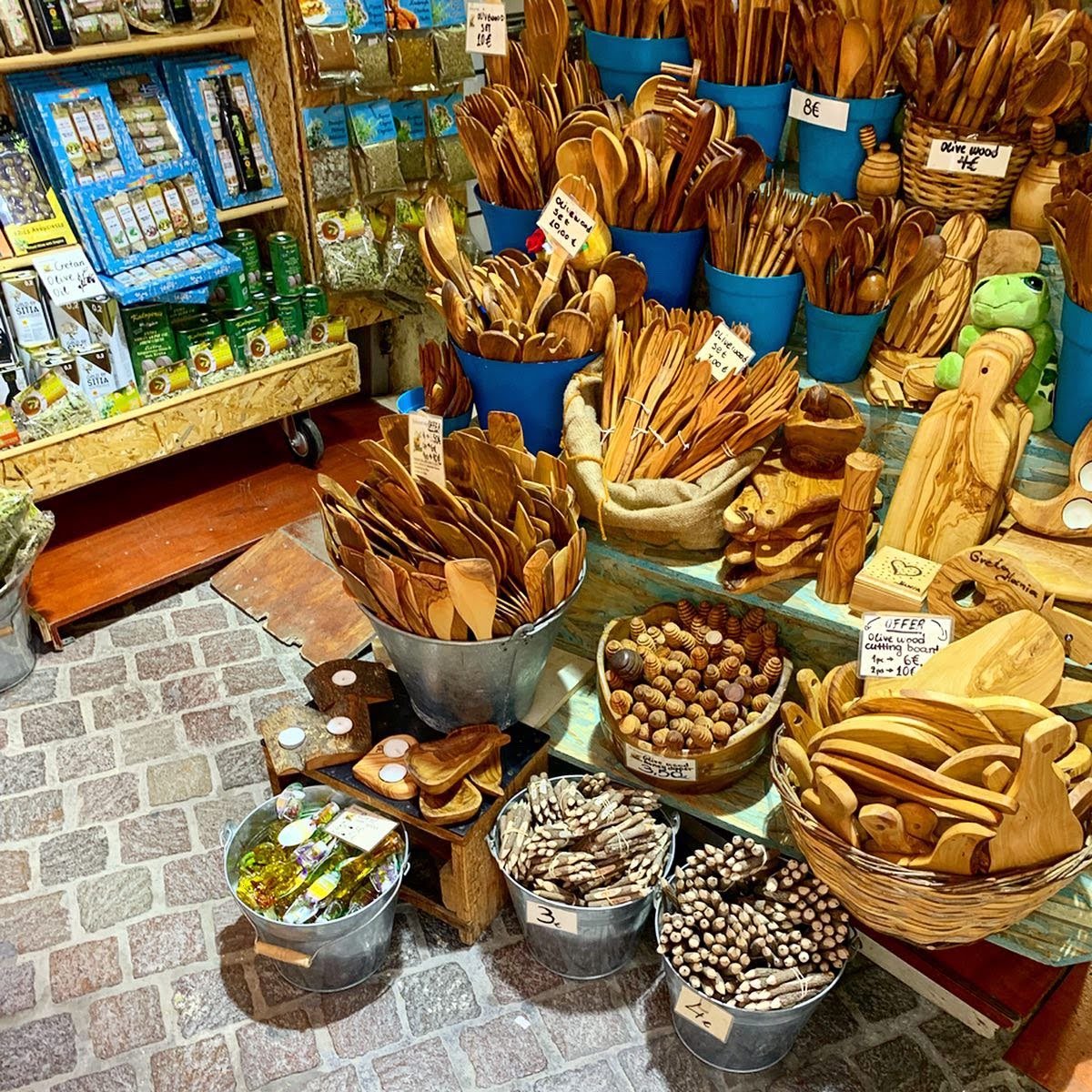 A market stall displaying a variety of wooden kitchen utensils including spoons, spatulas, and cutting boards. The items are organized in metal buckets and baskets. Shelves in the background hold packaged goods. The floor is cobblestone.