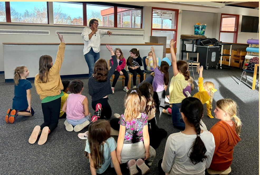 Kaylene standing in a classroom of children seated on the floor, some raising their hands.