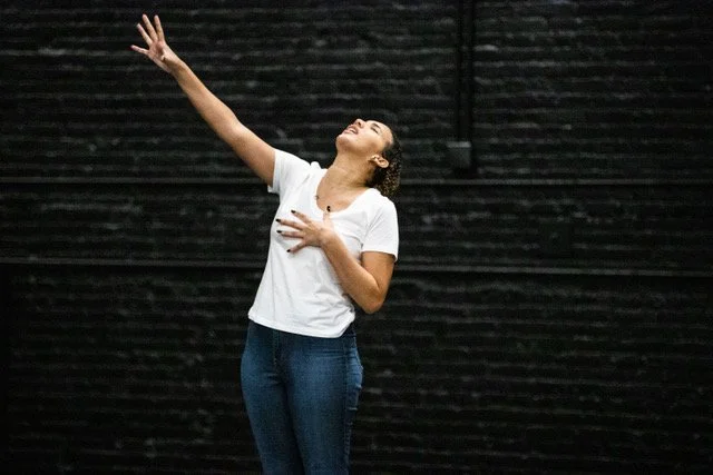 Kaylene in front of a black brick wall, left hand to her chest, face raised upward and right hand reaching up and out.