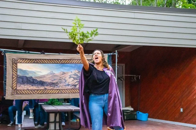 Kaylene wearing a purple cape over her shoulders while holding a green leafy branch upward. She is in an outdoor setting, with a framed image of mountains behind her.
