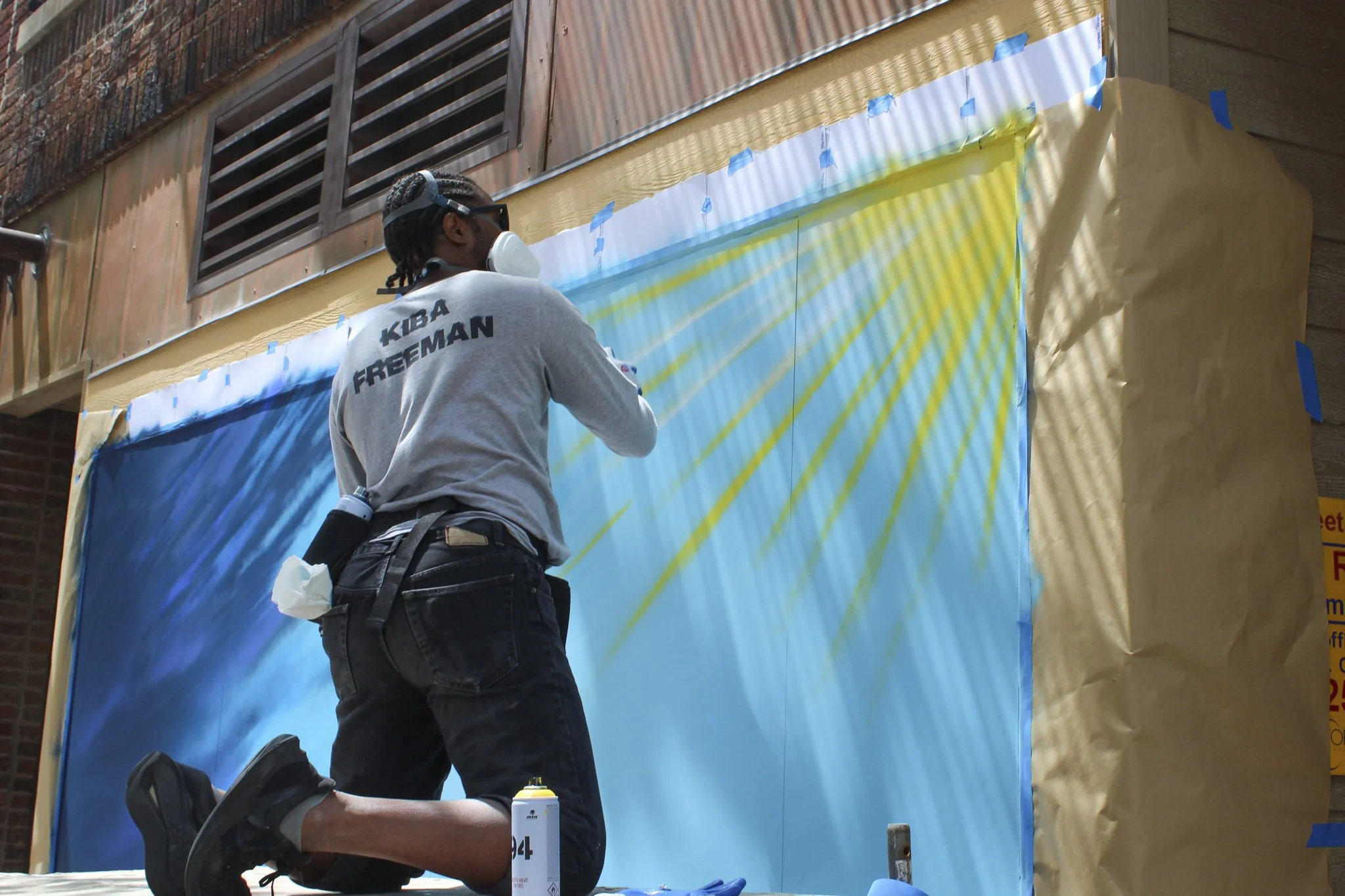 Kiba kneeling facing a mural-in-progress with blue background and sun in the upper right corner.