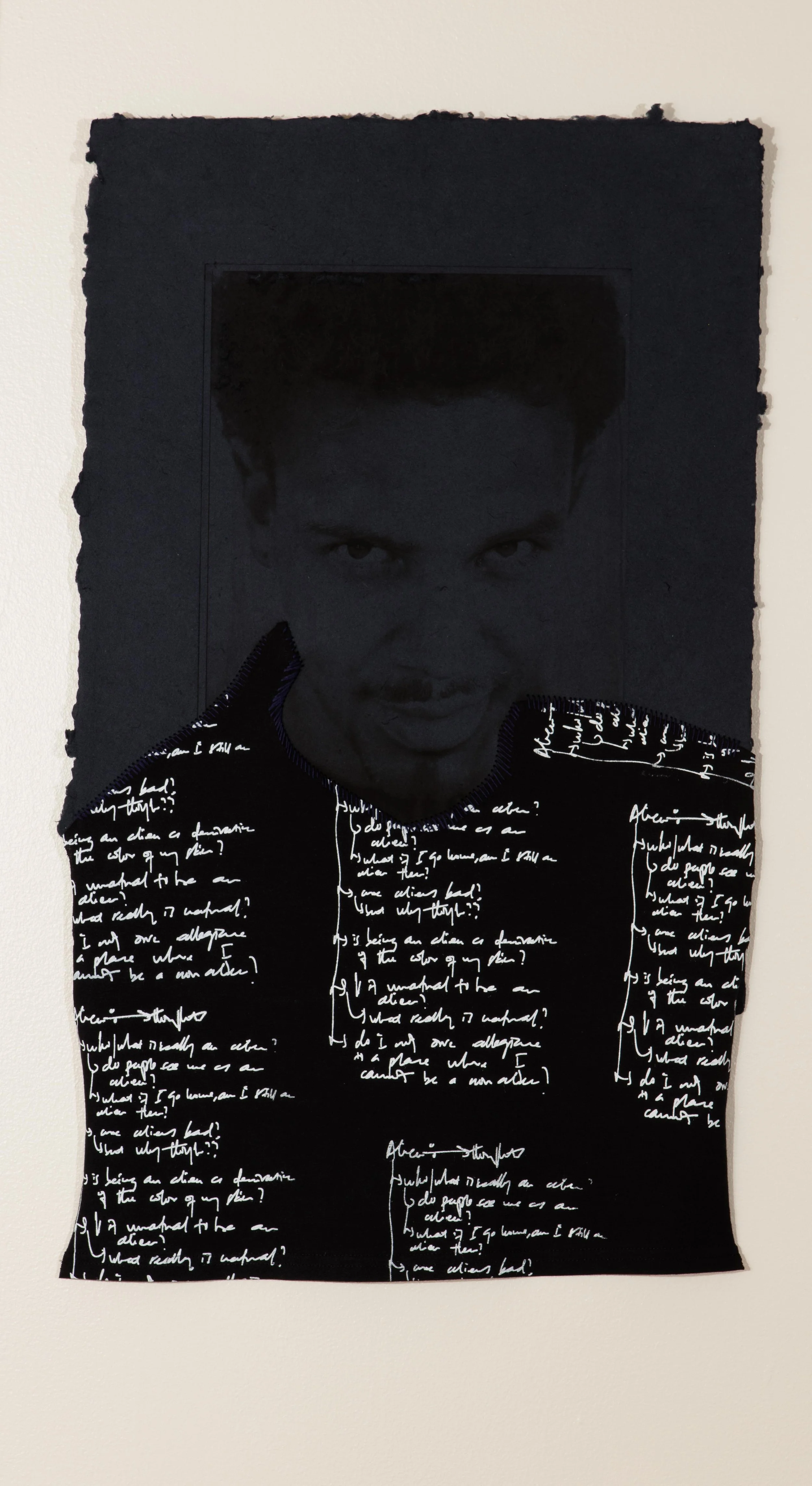 Portrait of a man with short hair and facial hair on dark gray paper. On the man's shirt is white handwritten text.