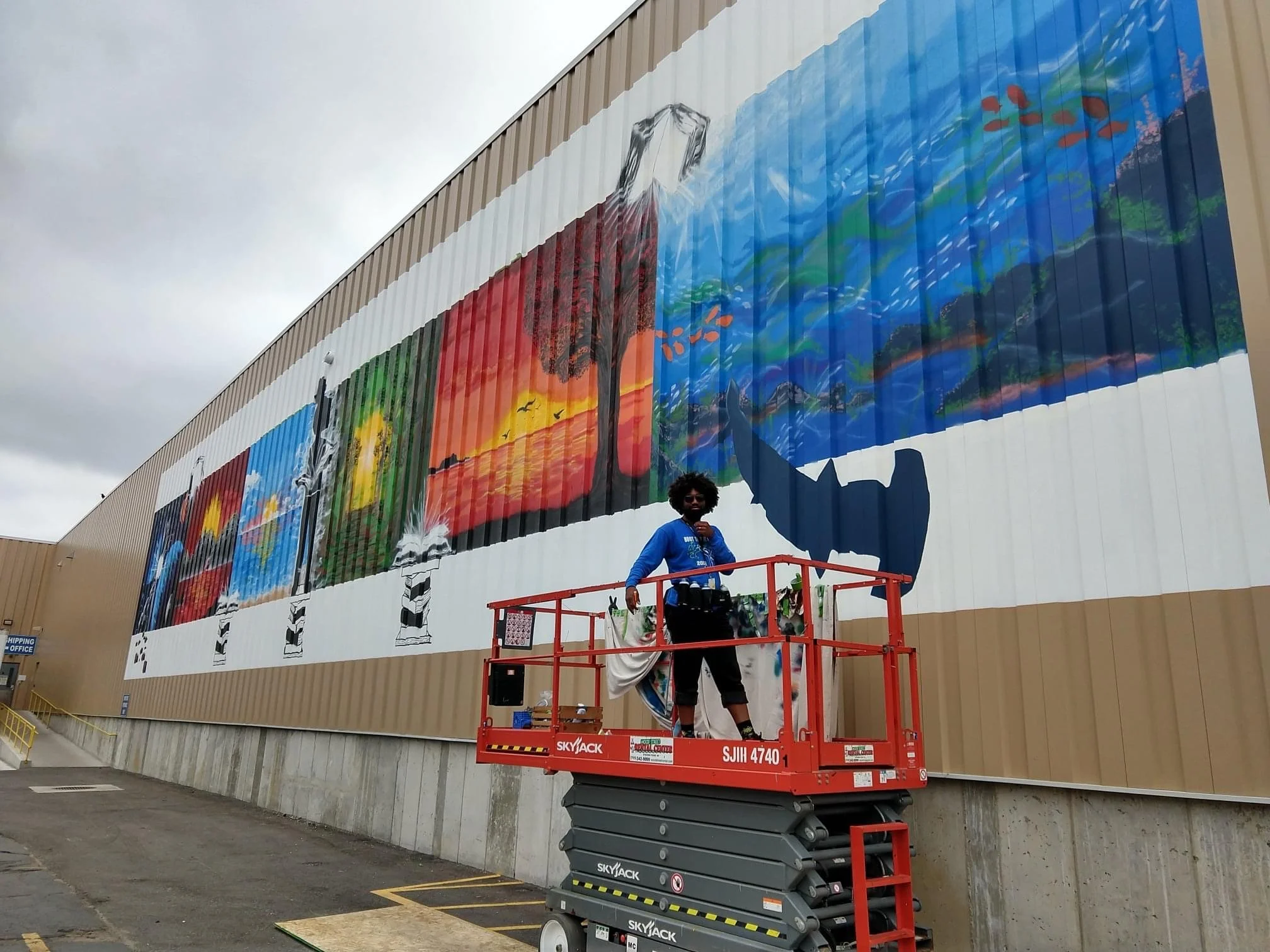 Kiba faces the camera, standing on a lift, with a large mural depicting seasons and natural landscapes.