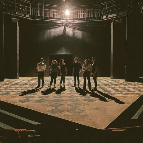 A bright light in the top center of the image, casting long shadows on six people standing on a checkered stage. Kaylene second from the right, hands crossed at her waist.