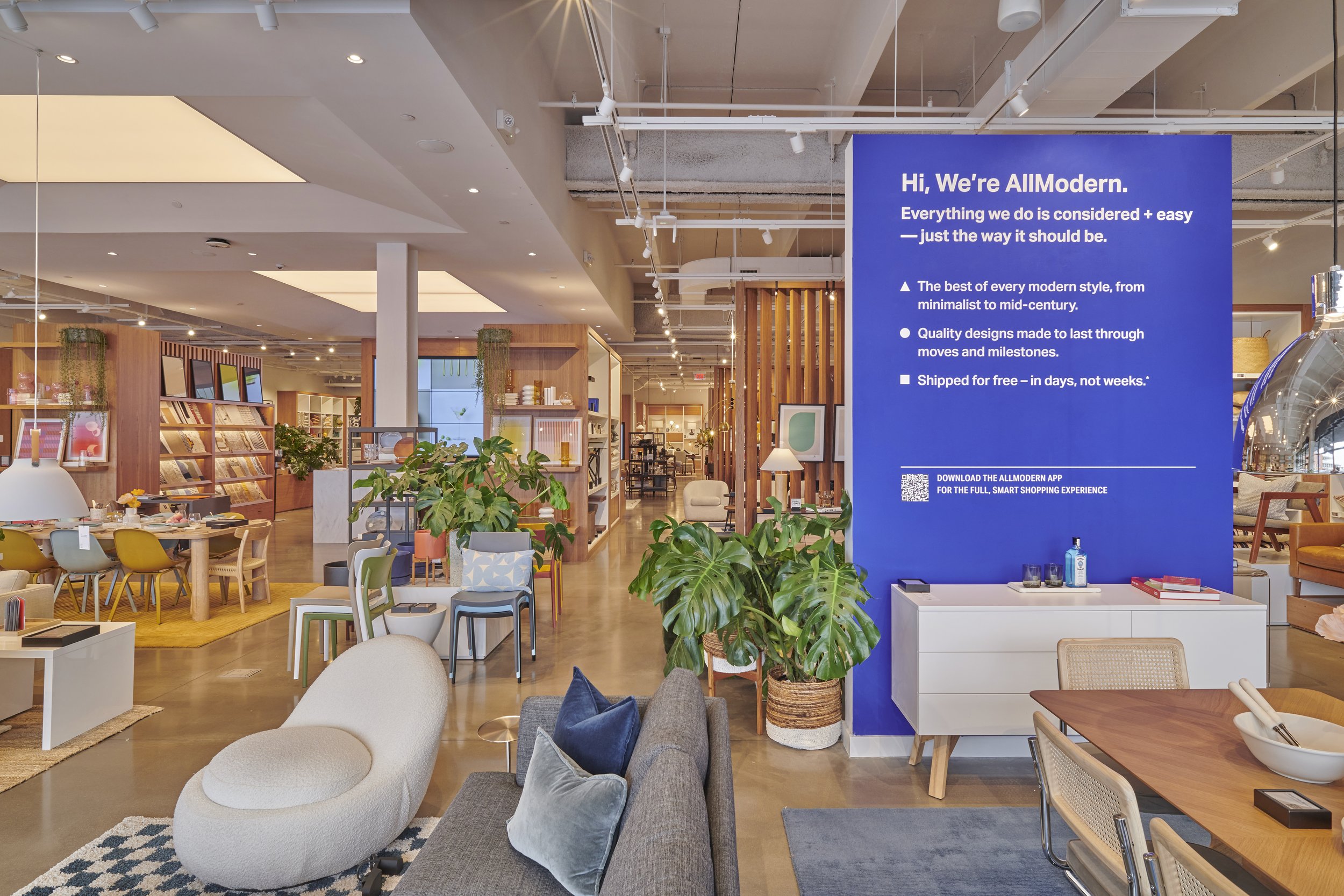 Modern furniture showroom interior featuring sofas, chairs, tables, and decor items. Large blue sign with store information on the right. Bright and open layout with plants and contemporary design elements.