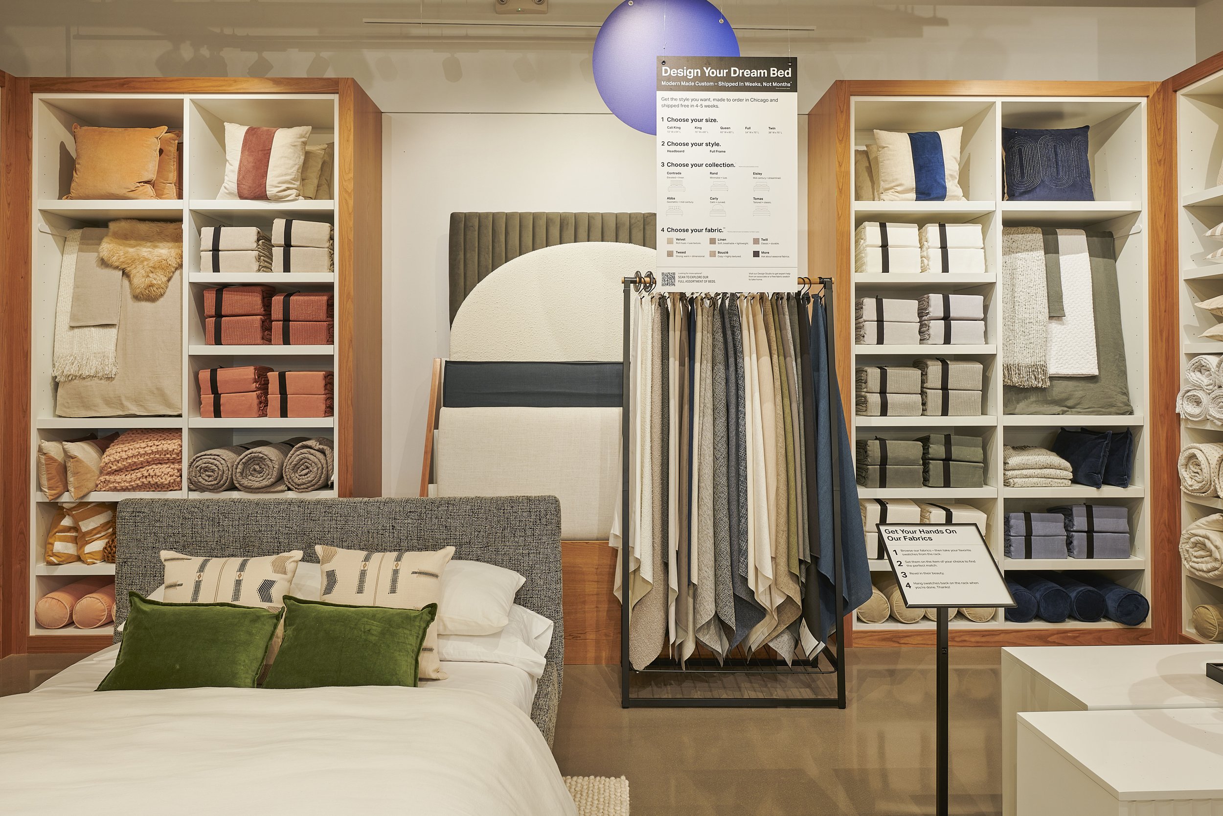 Bedroom showroom display with a bed, pillows, bedding fabrics, and organized shelves containing blankets and cushions. A sign provides customization options for designing a bed.