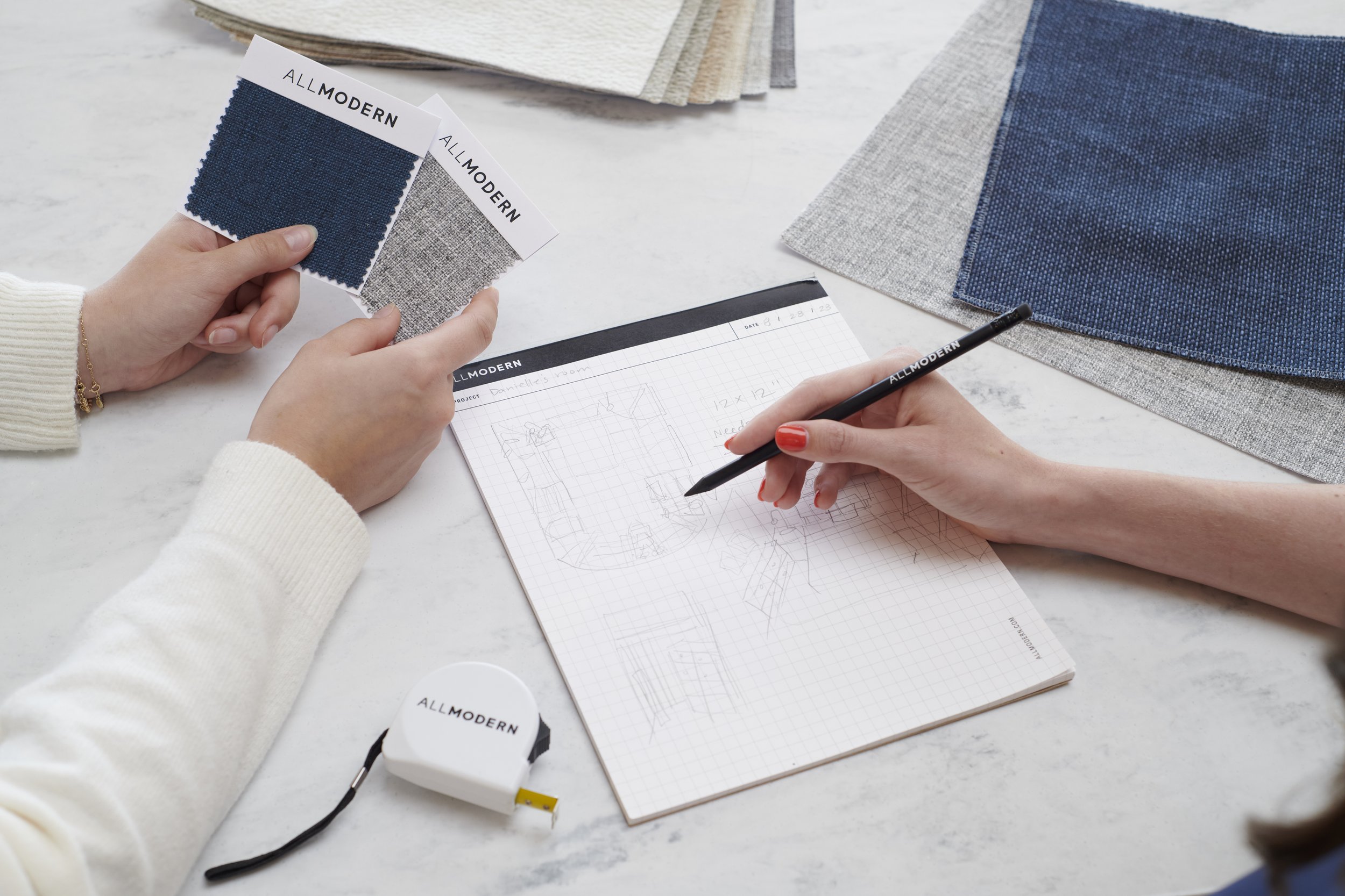 Two people working on a design project, with fabric swatches from AllModern, a notebook with room sketches, and a tape measure on a table.