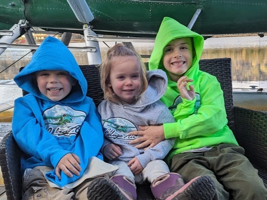 Three children wearing hoodies, sitting together and smiling in front of a seaplane.