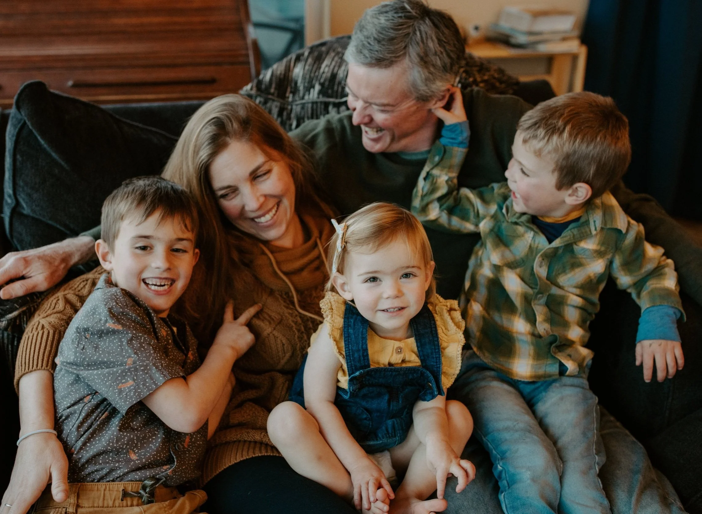 A smiling family sitting together on a couch, consisting of two adults and three young children, including two boys and a girl.