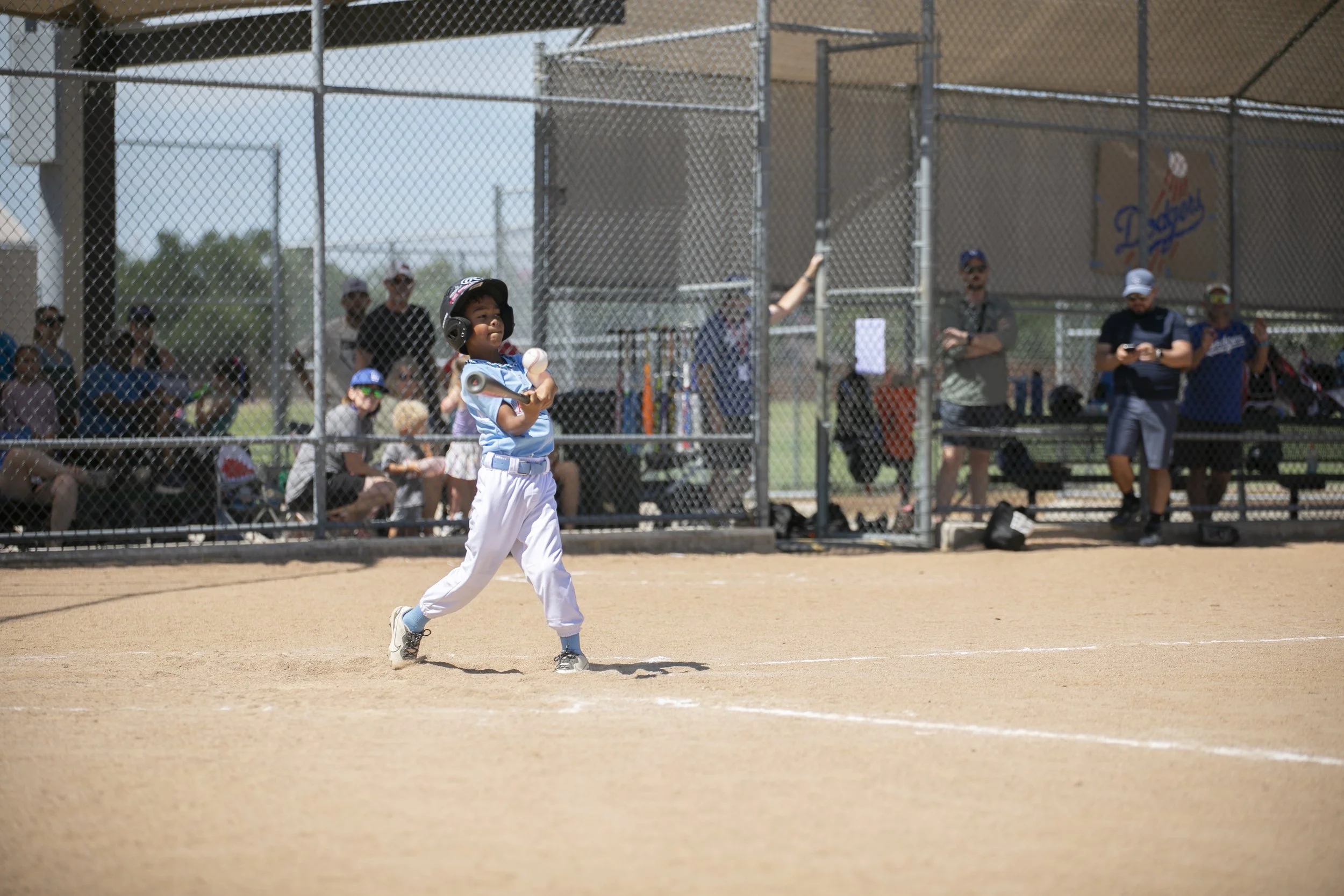 Young baseball player running on the field after hitting the ball during a game, with spectators watching from the dugout in the background.