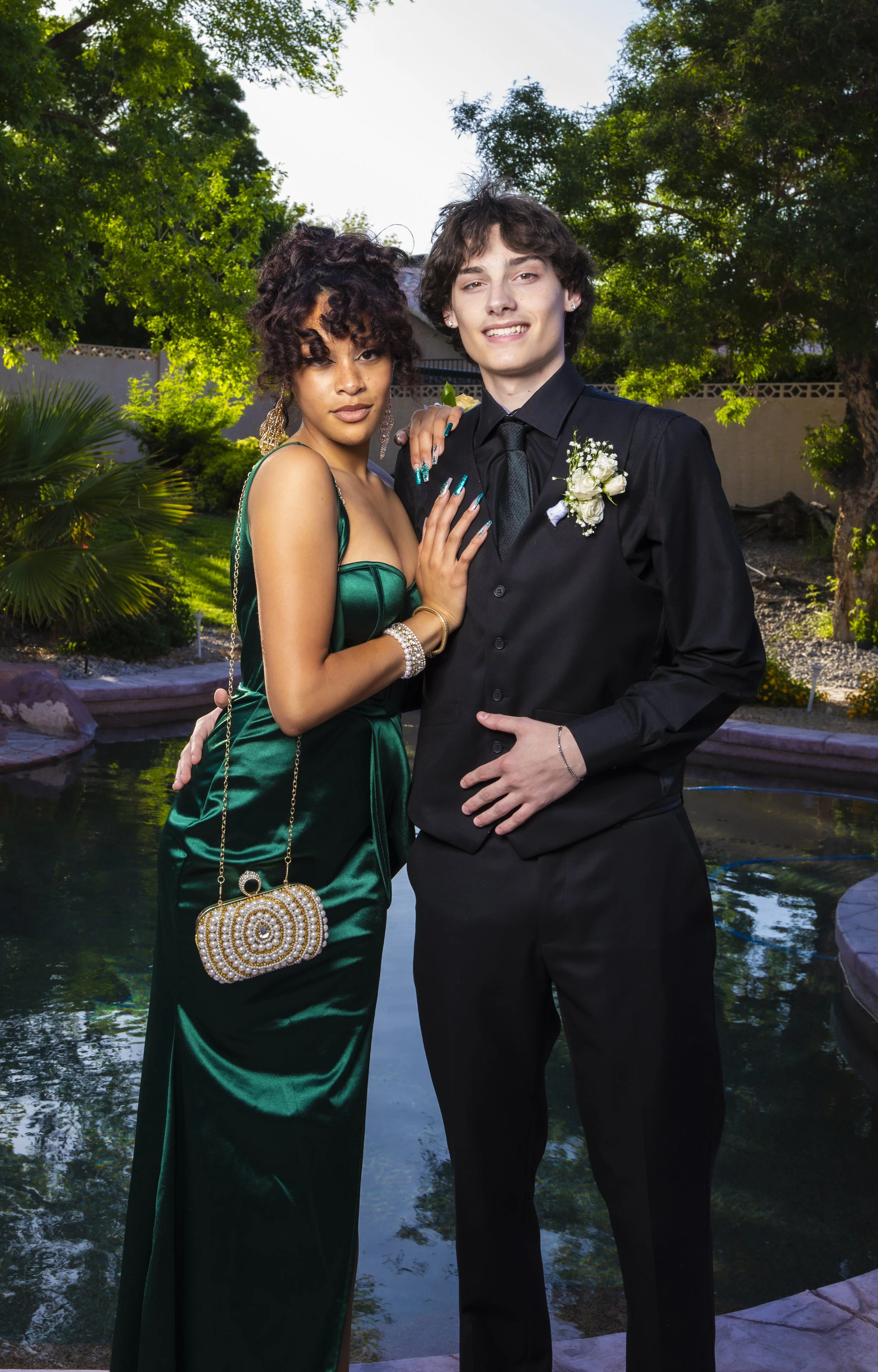 A young woman in a green satin gown and a young man in a black suit pose together outdoors near a pond, with trees and bushes in the background.