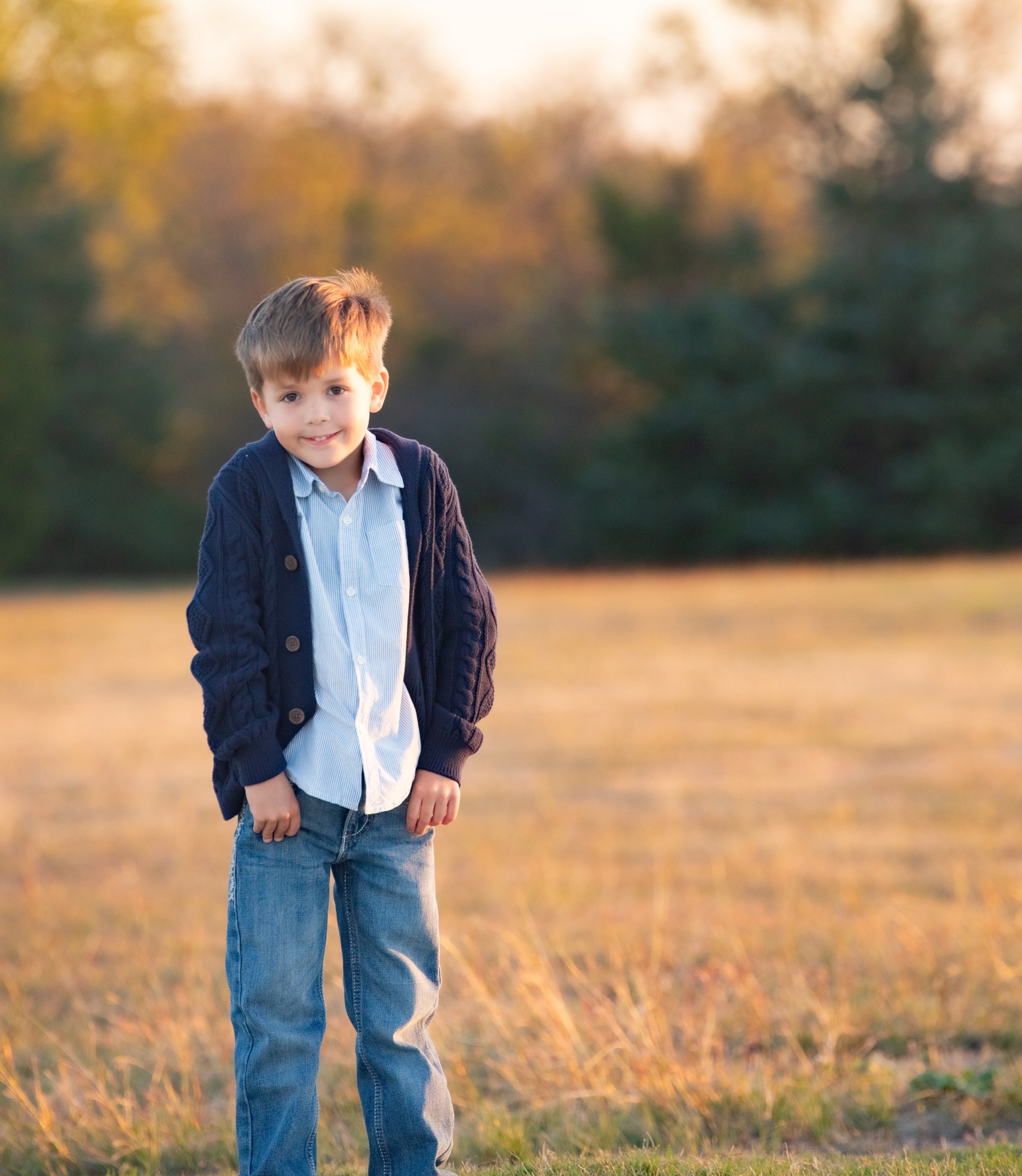 A young boy wearing a blue jacket, white shirt, and jeans standing in a grassy field during sunset.