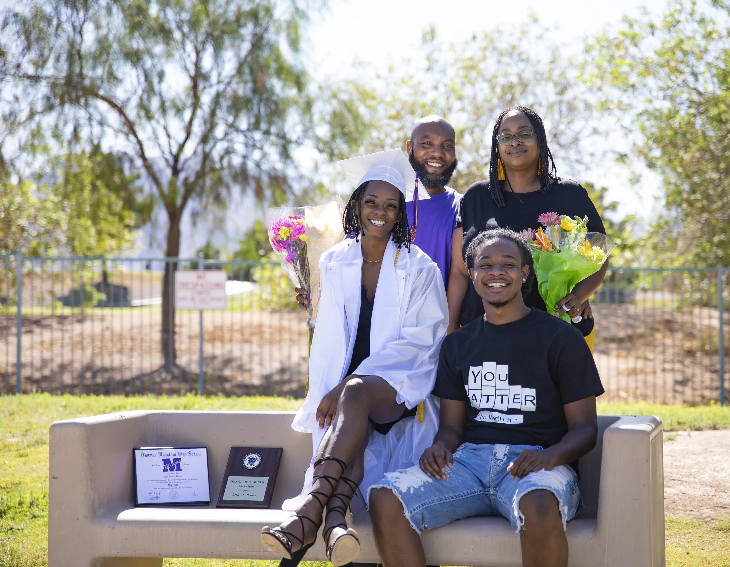 A group of five people celebrating outdoors, with a woman in a graduation cap and gown holding flowers, seated next to a young man in a black shirt with the words "You matter" on it, and others standing behind them holding bouquets of flowers. They a