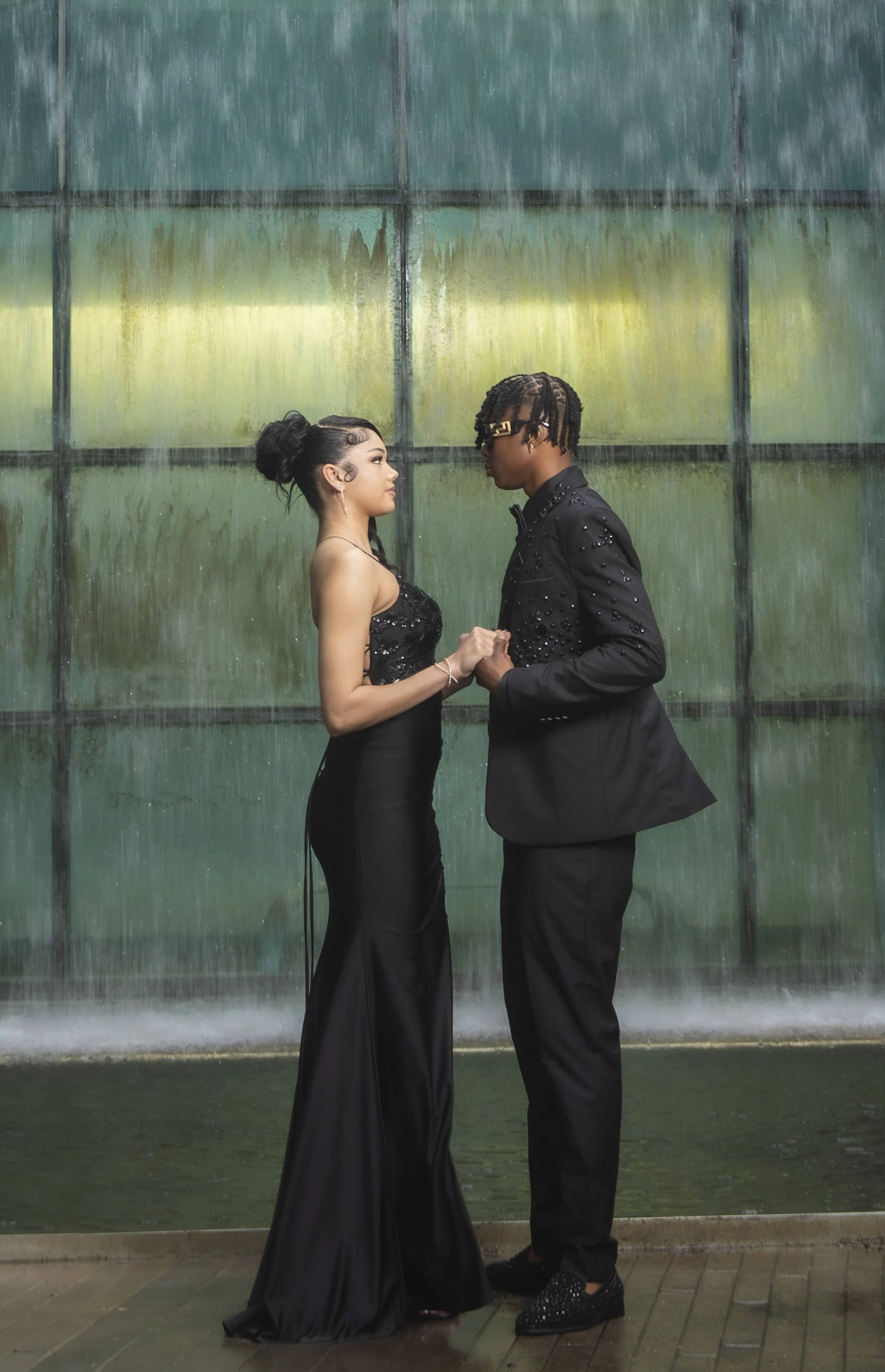 A man and woman dressed in black formal attire facing each other, holding hands, standing in front of a large water fountain wall.
