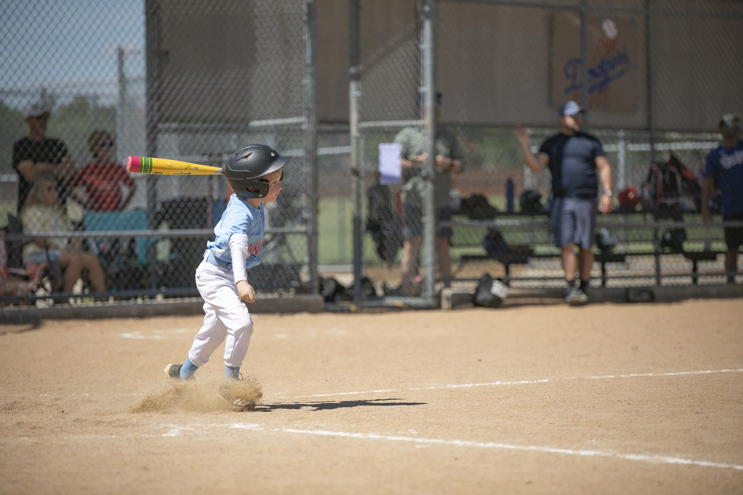 A young boy in a light blue shirt, white pants, and a black helmet running on a baseball field with a toy rocket launcher on his shoulder, kicking up dust as he runs.