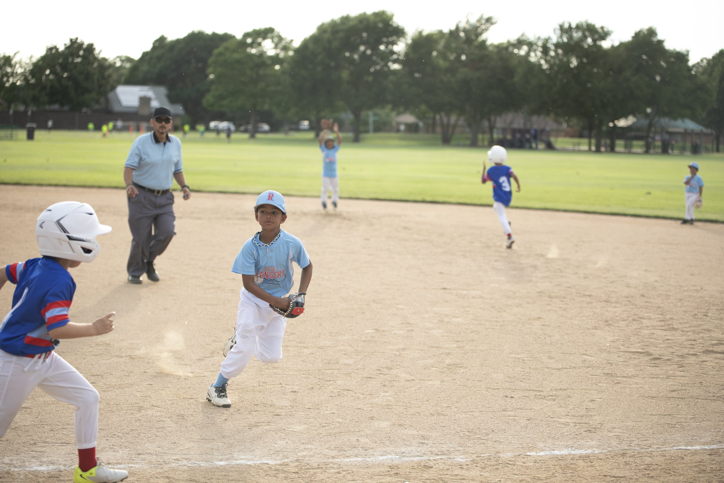 Children playing baseball with an umpire on a field, some children running and others waiting, in a park with trees and houses in the background.
