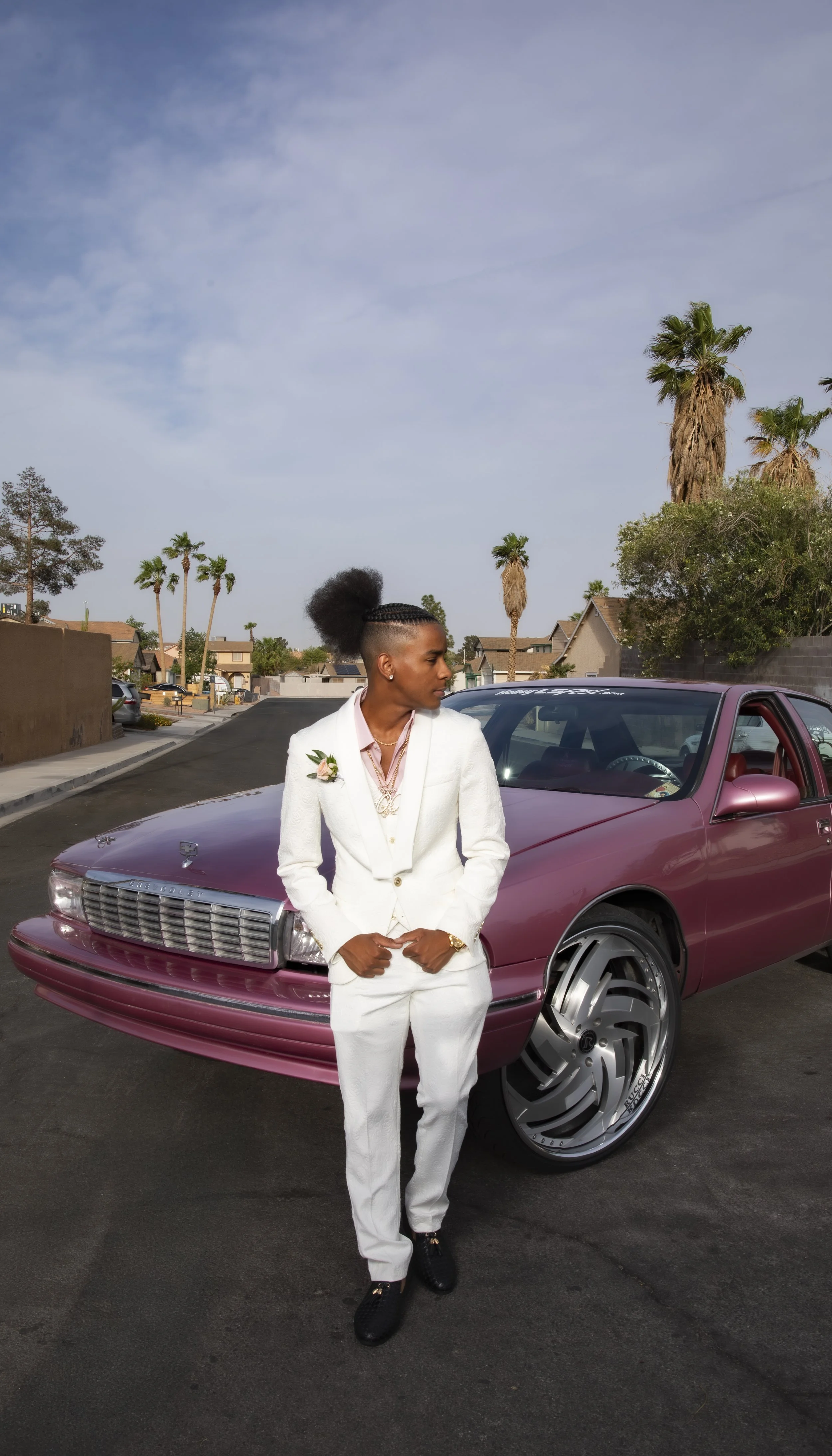 A woman in a white suit with a pink flower corsage and gold jewelry standing on the street in front of a pink luxury car with large rims, under a partly cloudy sky with palm trees in the background.