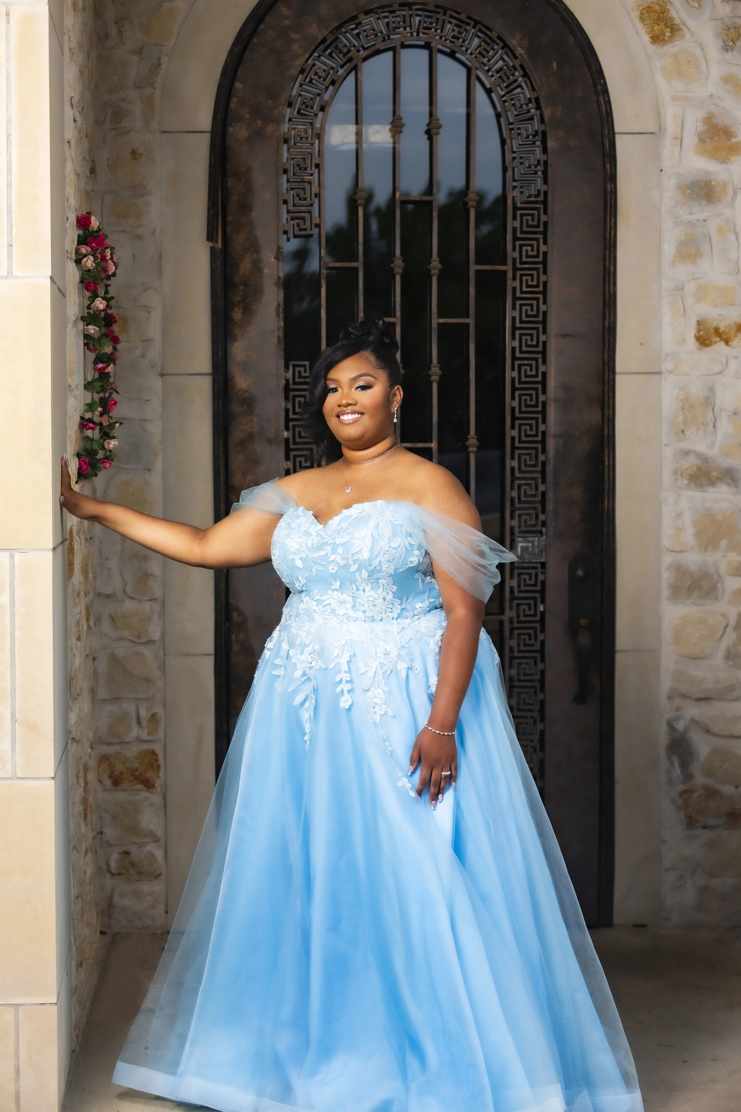 A woman in a blue formal gown standing near a stone wall and a black metal gate, smiling at the camera.