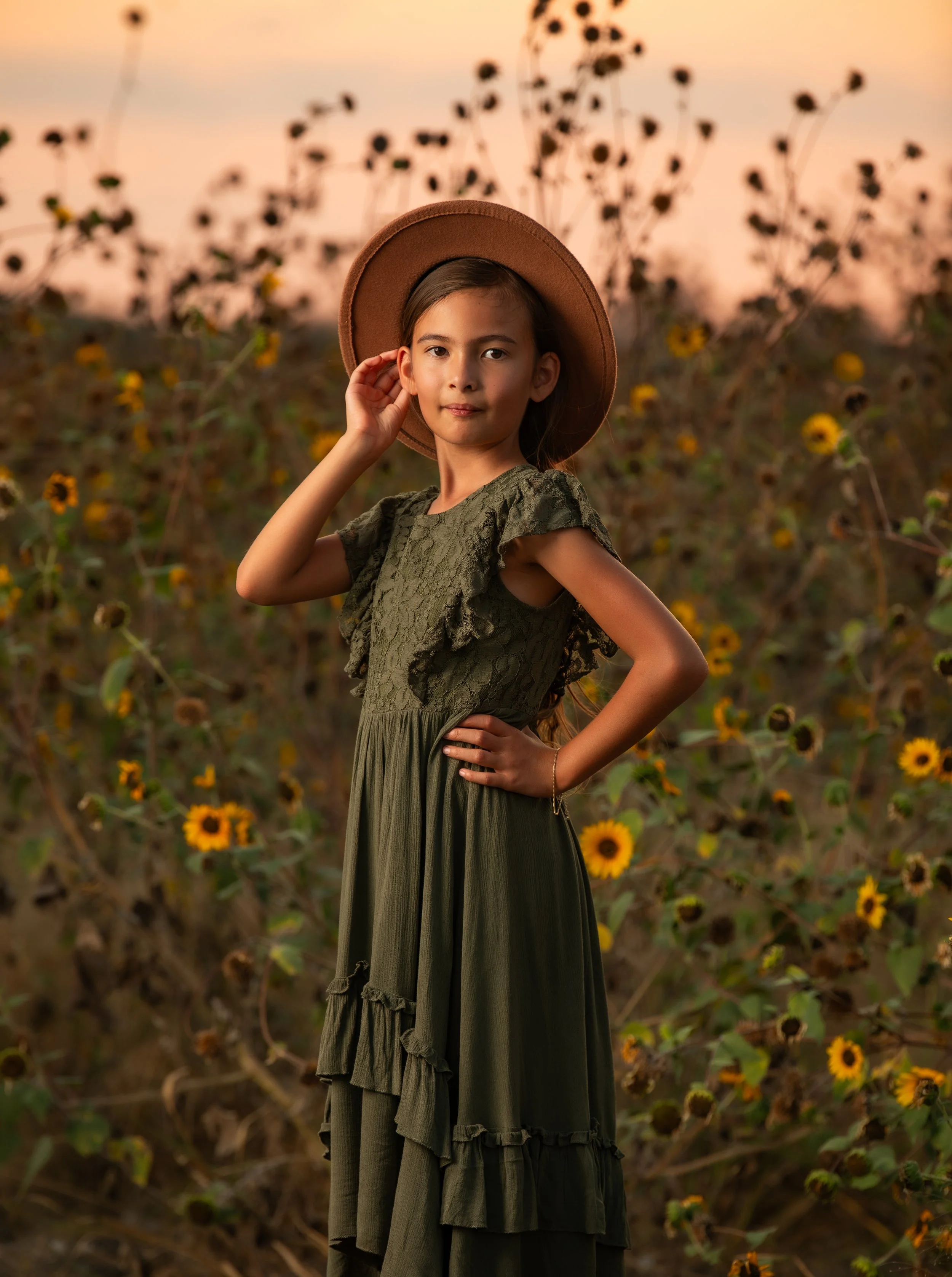 A girl in a green dress and wide-brimmed hat stands in a sunflower field during sunset.