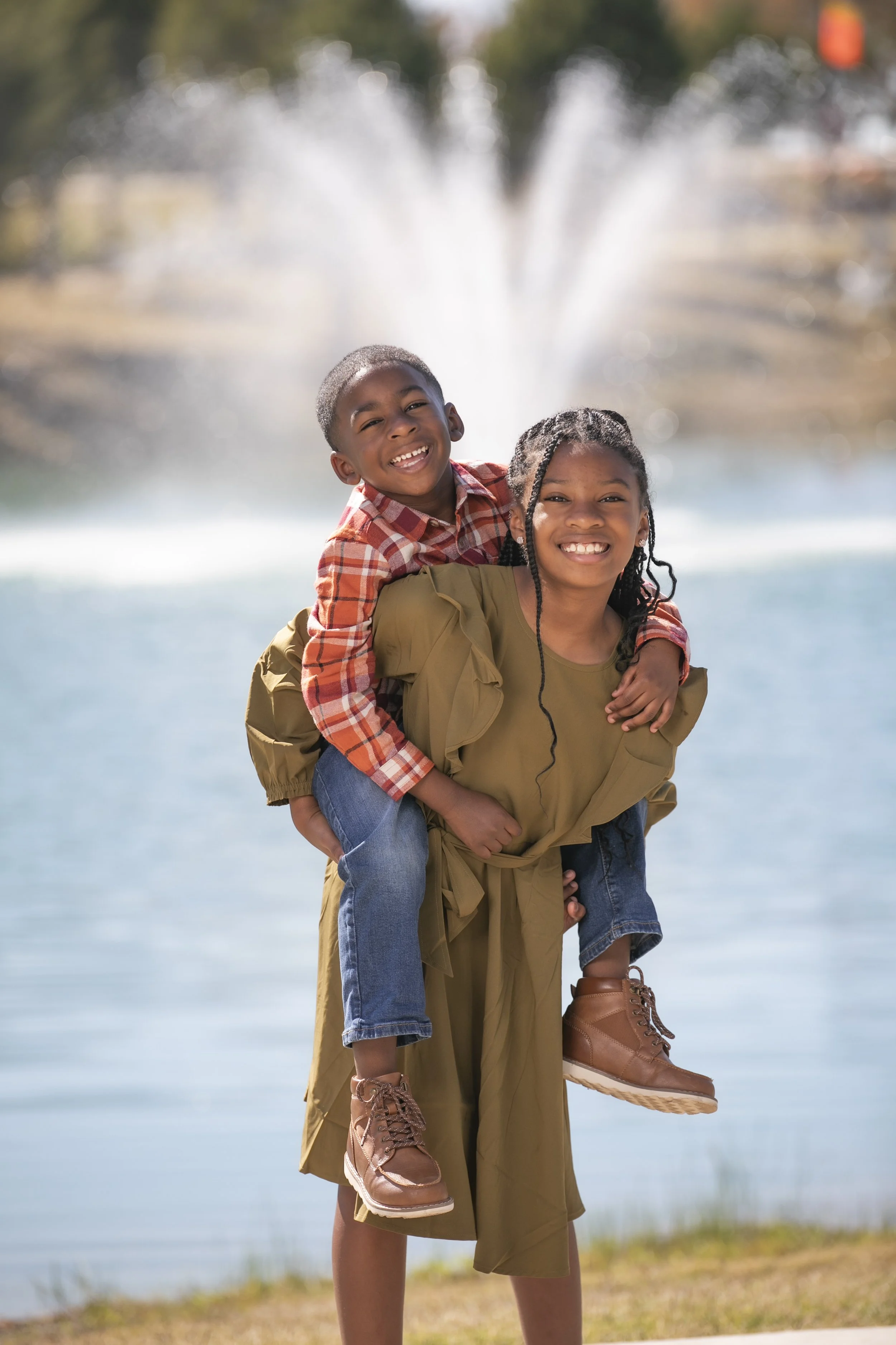 Two children, a girl and a boy, smiling and having fun outdoors near a fountain at the courthouse in Princeton Tx, with the boy giving the girl a piggyback ride.