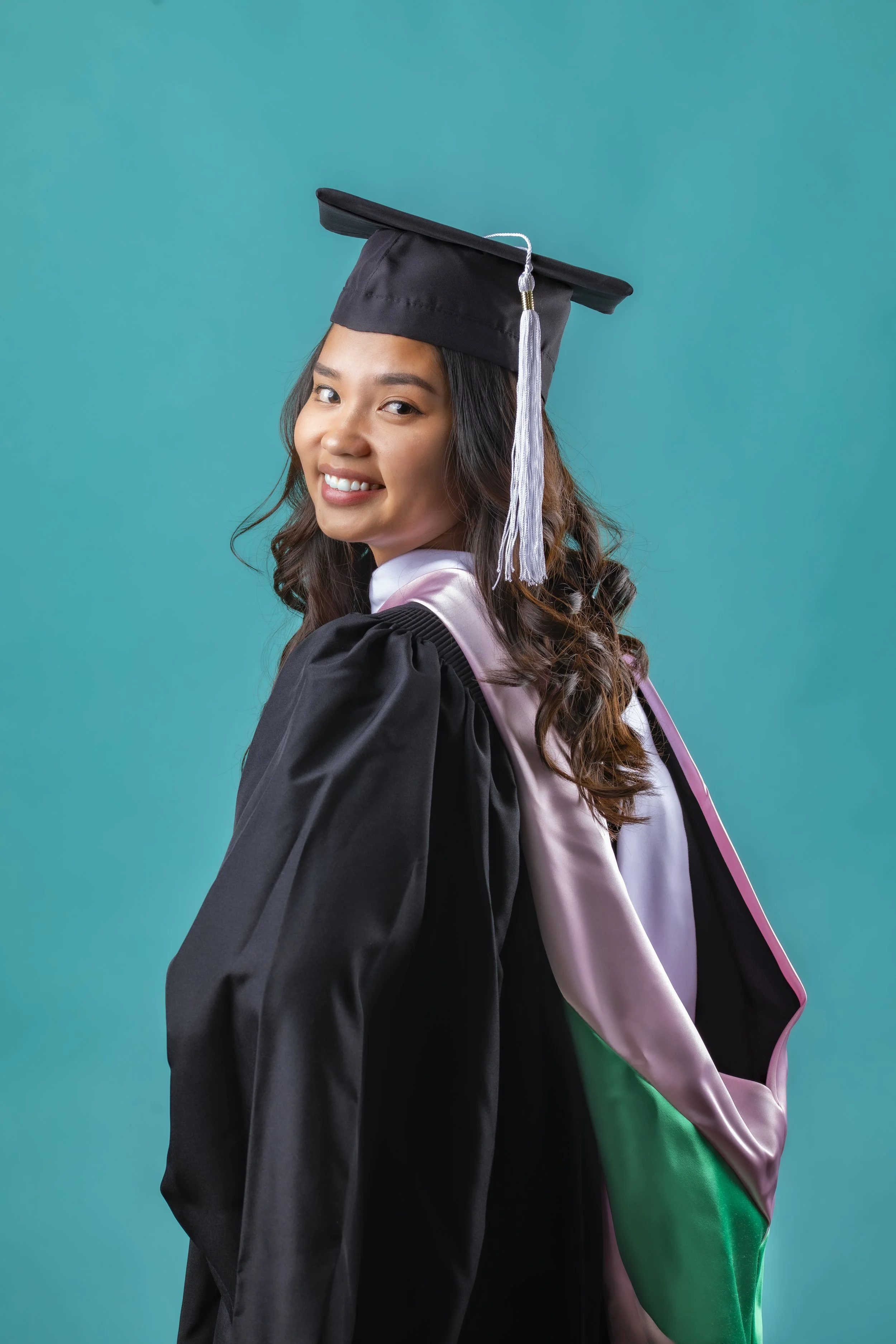 A young woman in graduation attire, including a black cap and gown, with a colorful hood, smiling at the camera against a teal background.