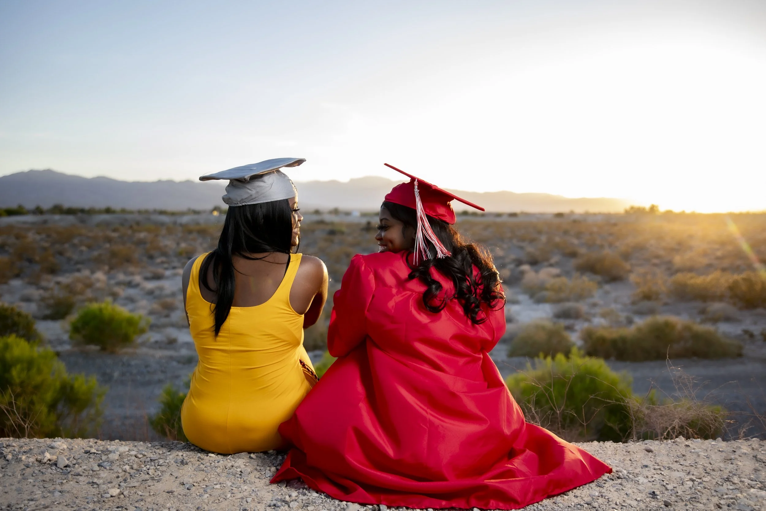 Two women sitting on the ground in a desert landscape at sunset, one in a yellow dress with a white cap on her head and the other in a red graduation gown with a matching cap, looking at each other.