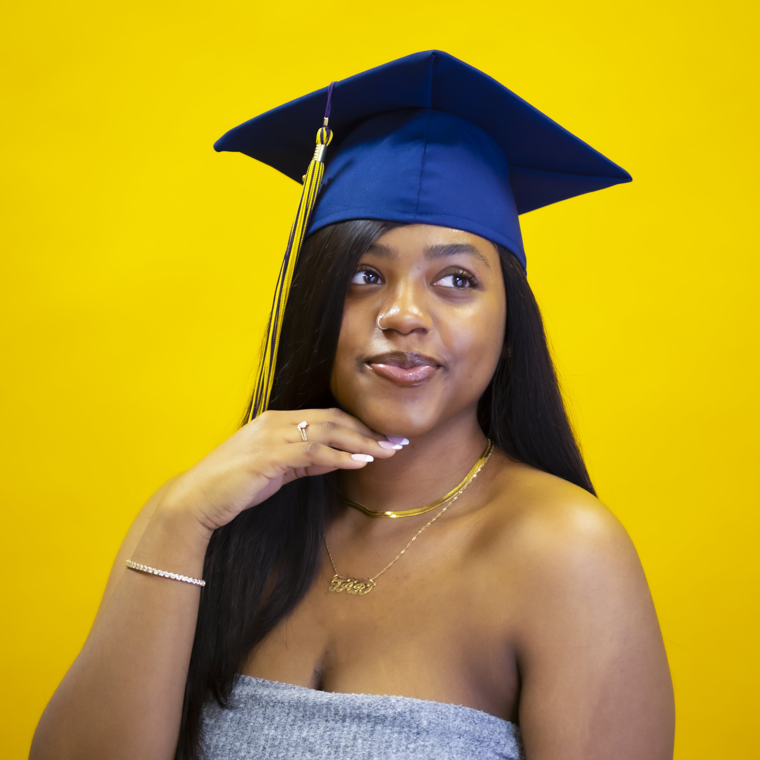 A young woman wearing a blue graduation cap with a yellow and purple tassel, smiling against a yellow background.