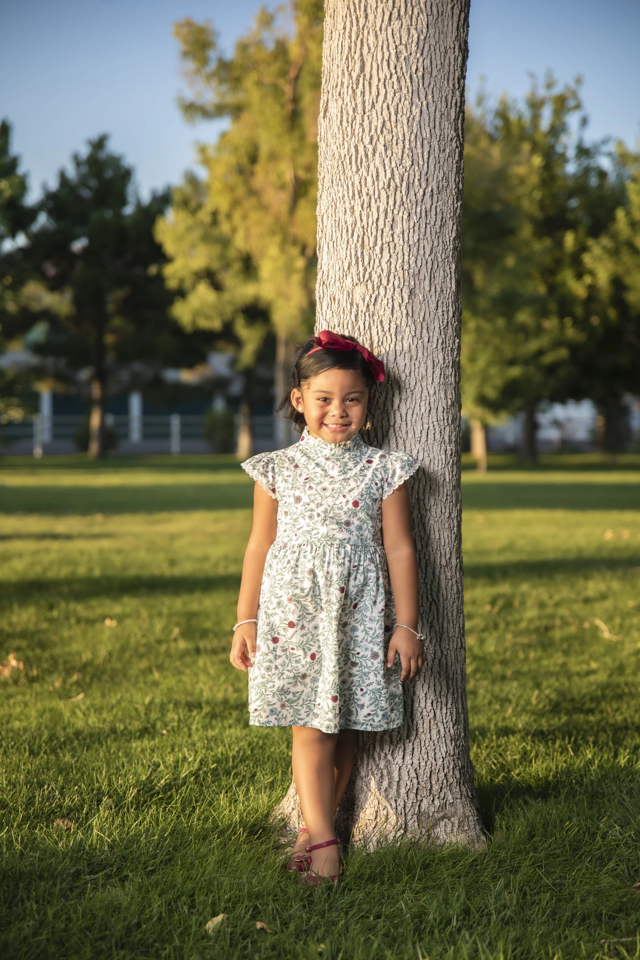 A young girl with dark hair in a dress standing next to a tree in a park during sunset.