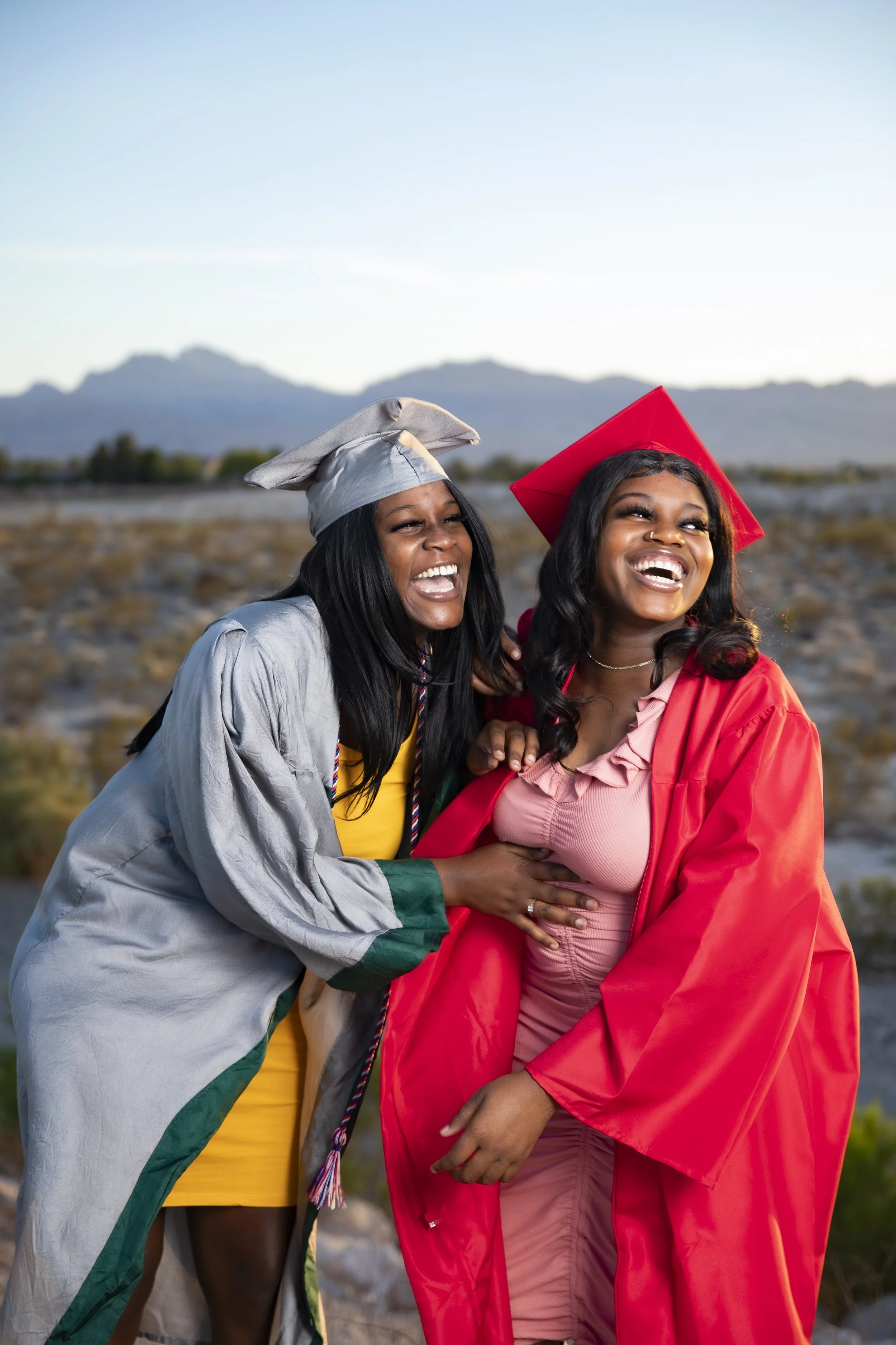 Two women celebrating graduation outdoors, one in a gray graduation gown and cap, the other in a red cap and gown, both smiling and laughing.