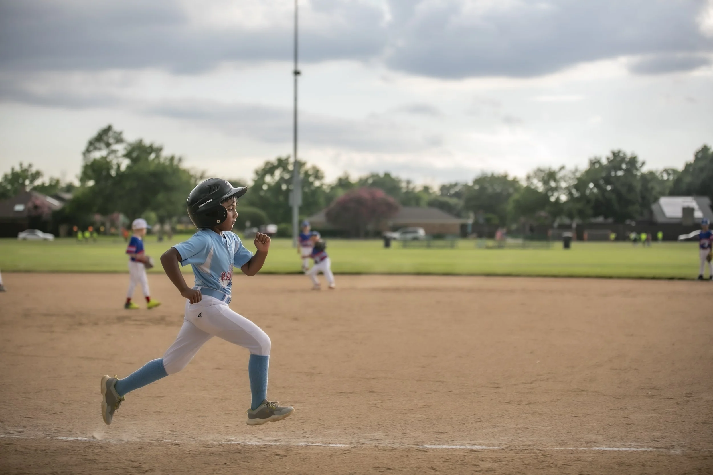 A young baseball player in a black helmet and light blue uniform running on the field during a game, with other players and a grassy area with trees in the background.