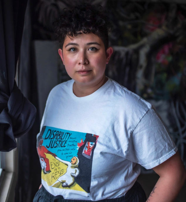 Élaina is a light-skinned Filipinx femme with short dark curly hair. She is posing seriously while wearing a Sins Invalid tshirt.
