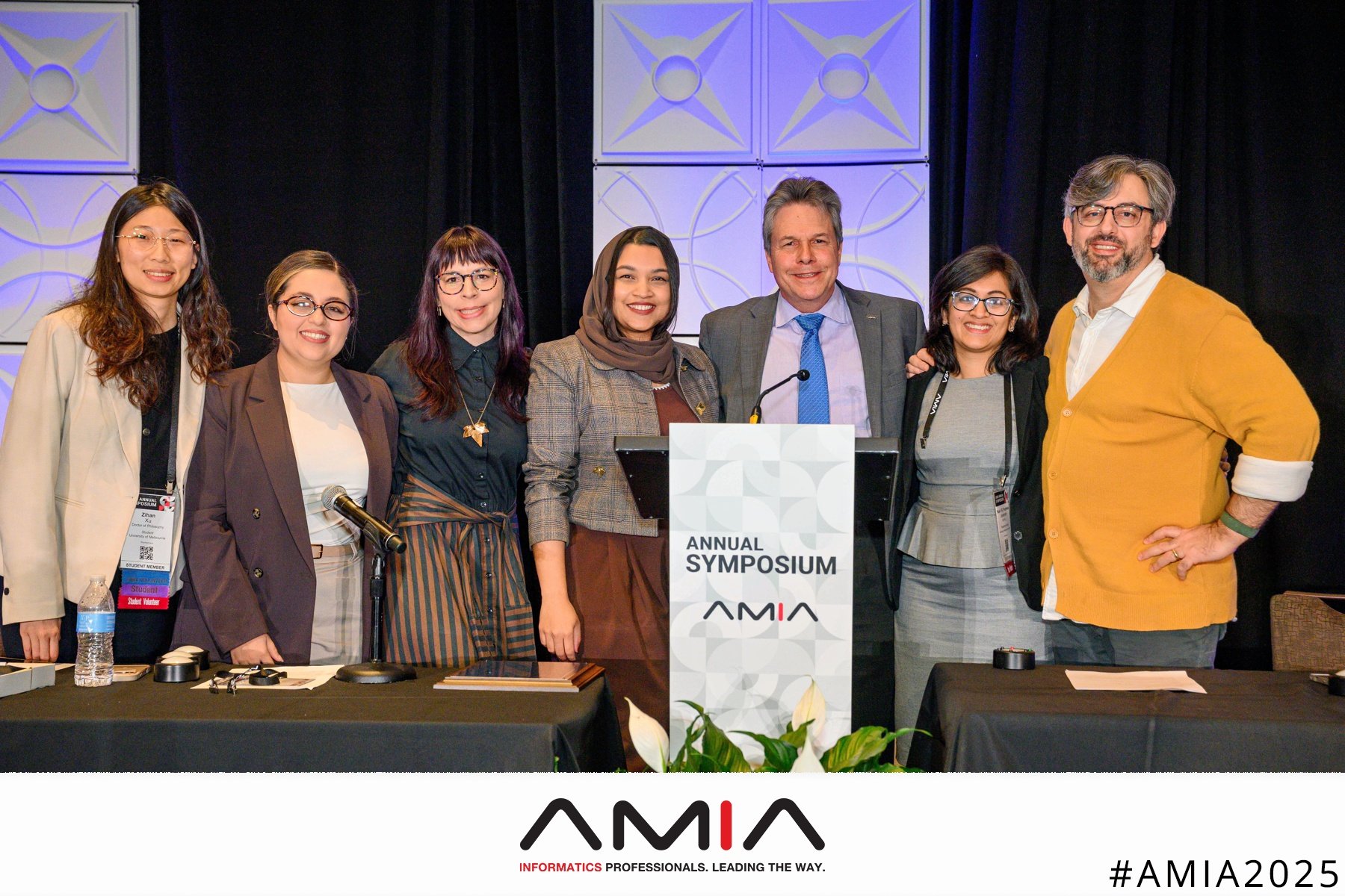 Seven people, including Malin Britt Lalich, smiling at a podium that reads "Annual Symposium AMIA" after the Informatics Year in Review Closing Keynote.