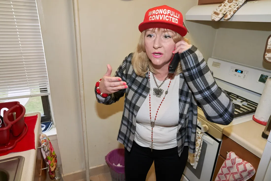 A woman with blonde hair talking on a cell phone in a kitchen, wearing a red hat that says 'Wrongfully Convicted' and a black plaid blazer.