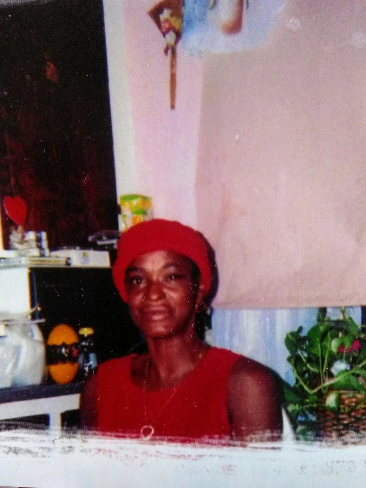 A woman wearing a red top and red hat sitting in a kitchen with a fridge, kitchen supplies, and a plant in the background.