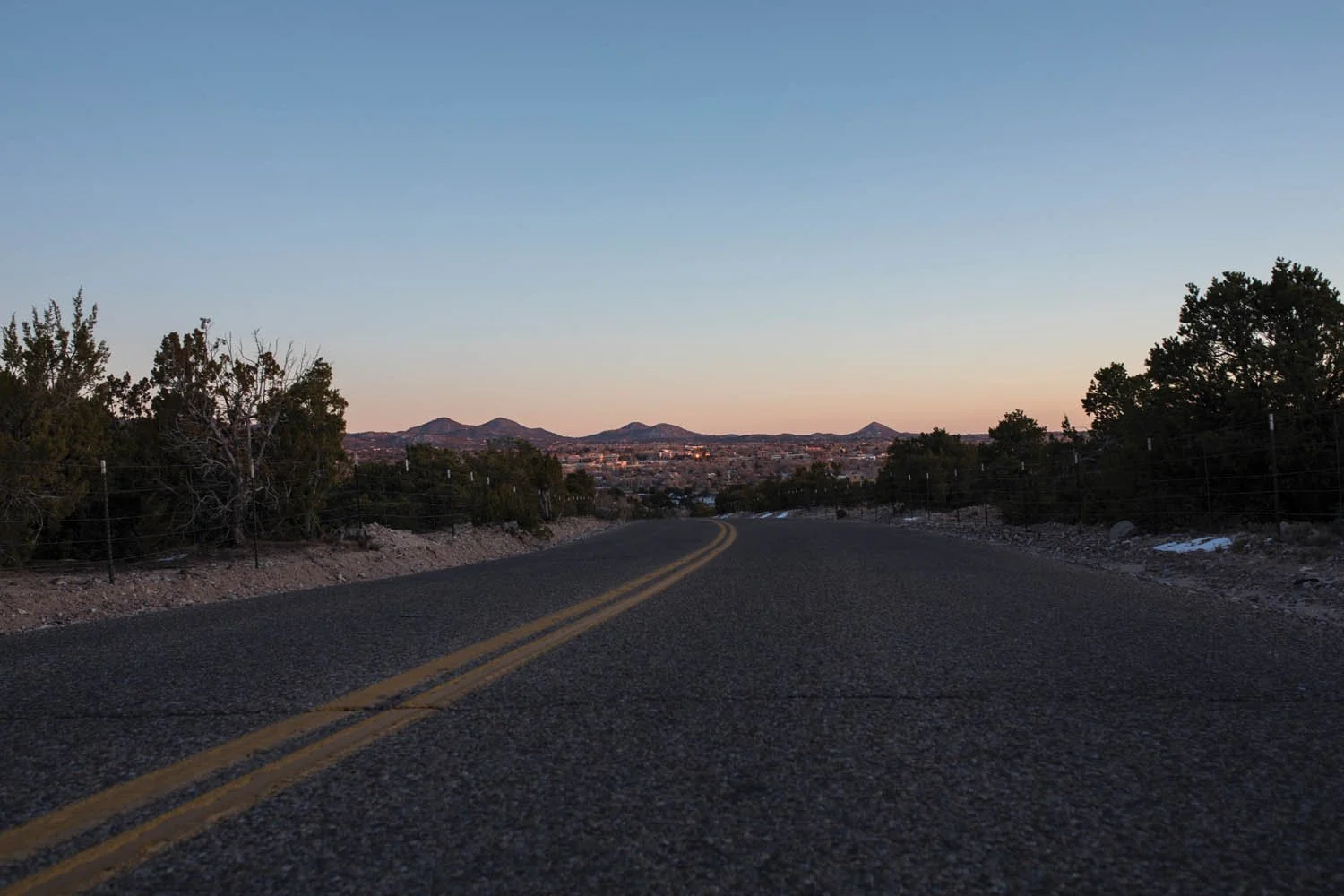 A rural asphalt road with yellow double lines curves into the distance, flanked by desert shrubs and trees, with mountains in the background and a clear evening sky.