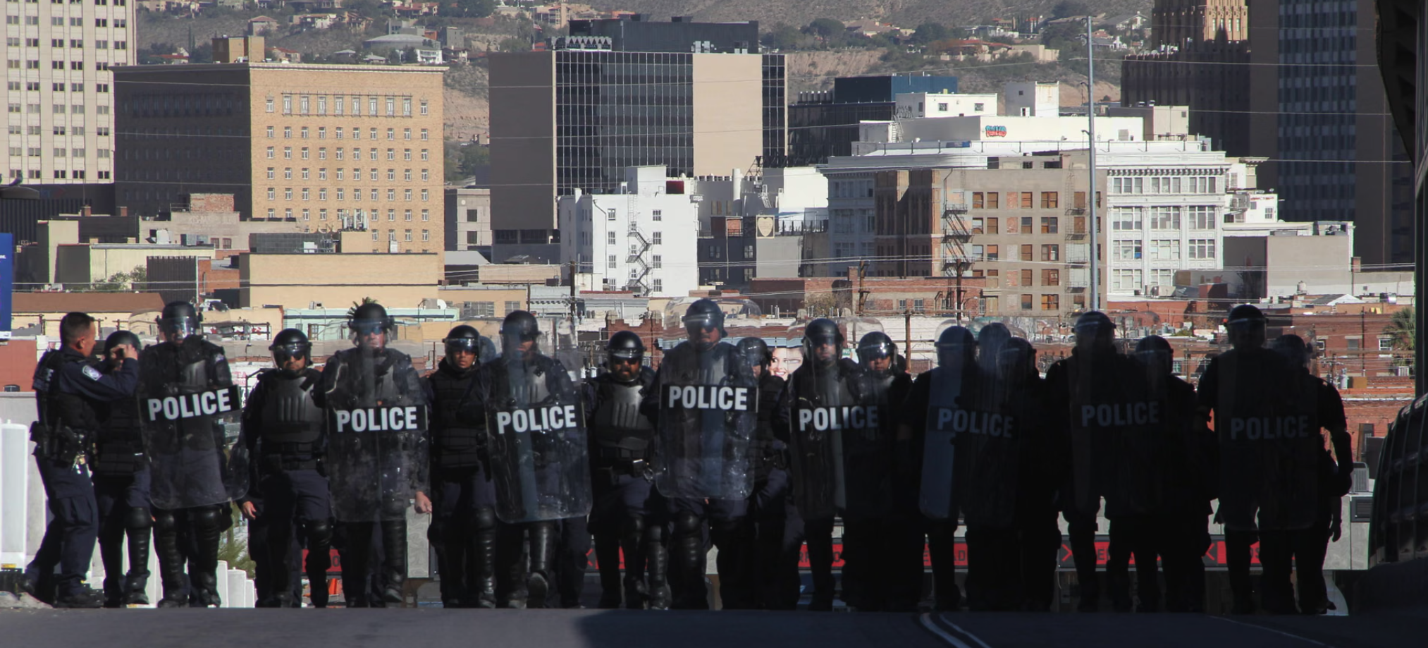 Multiple police officers in riot gear standing in a line, holding shields labeled 'POLICE', with a city skyline in the background.