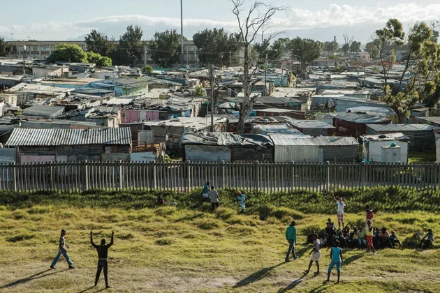 Children playing outdoors in a grassy area with a fence behind them, overlooking a large makeshift settlement with scattered tin-roofed structures in the background.