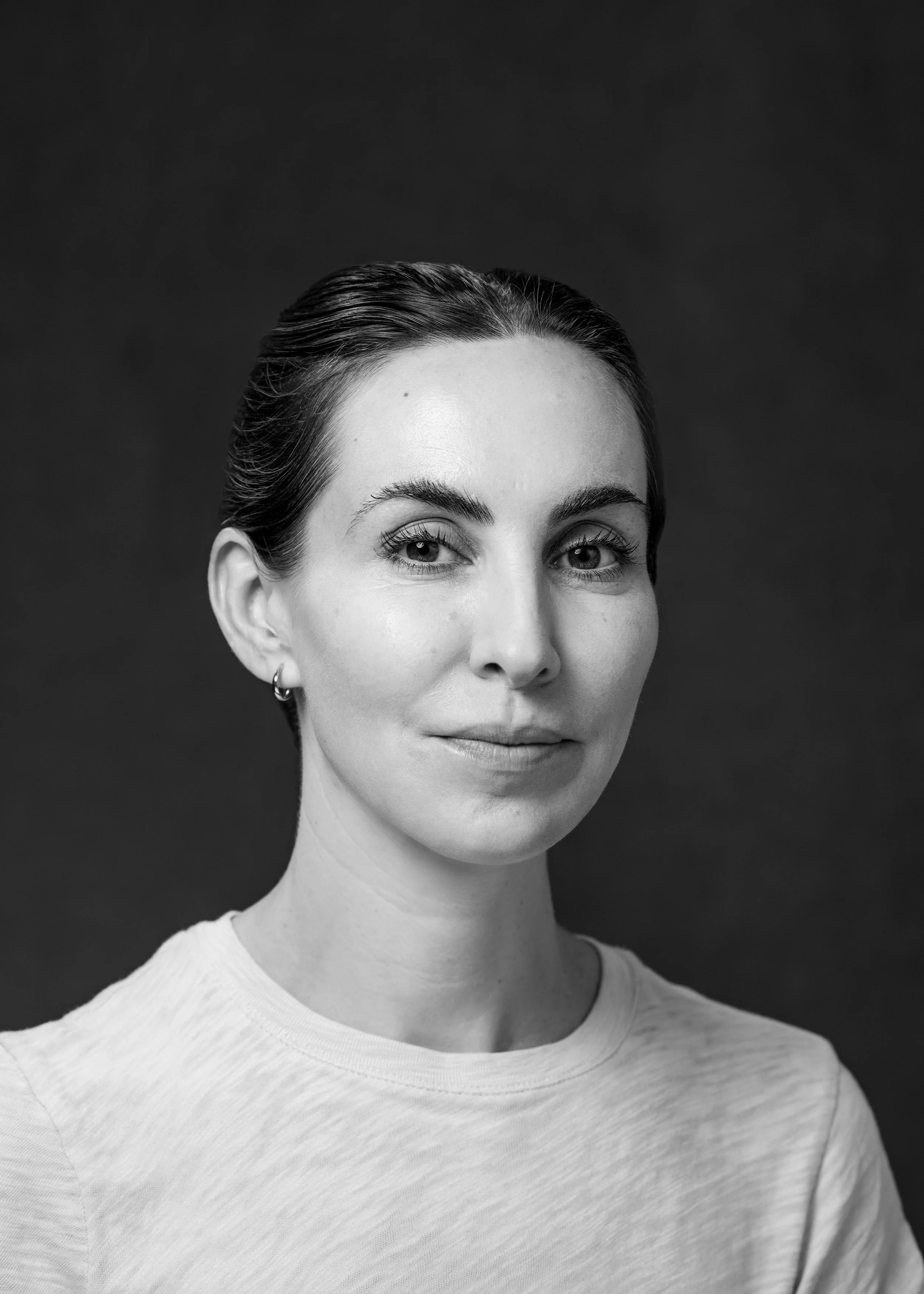 Black and white portrait of a woman with dark hair pulled back, wearing a light-colored shirt and hoop earrings, against a dark background.