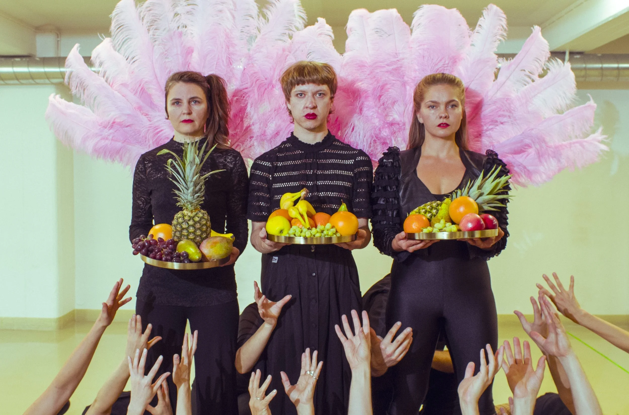 Kolme naista seisoo suorassa, kätensä ojennettuina eteenpäin, jokainen pitää makeakori, jossa on erilaisia hedelmiä, kuten ananas, banaaneja ja appelsiineja. Taustalla on suuri vaaleanpunainen sulka-asetelma.