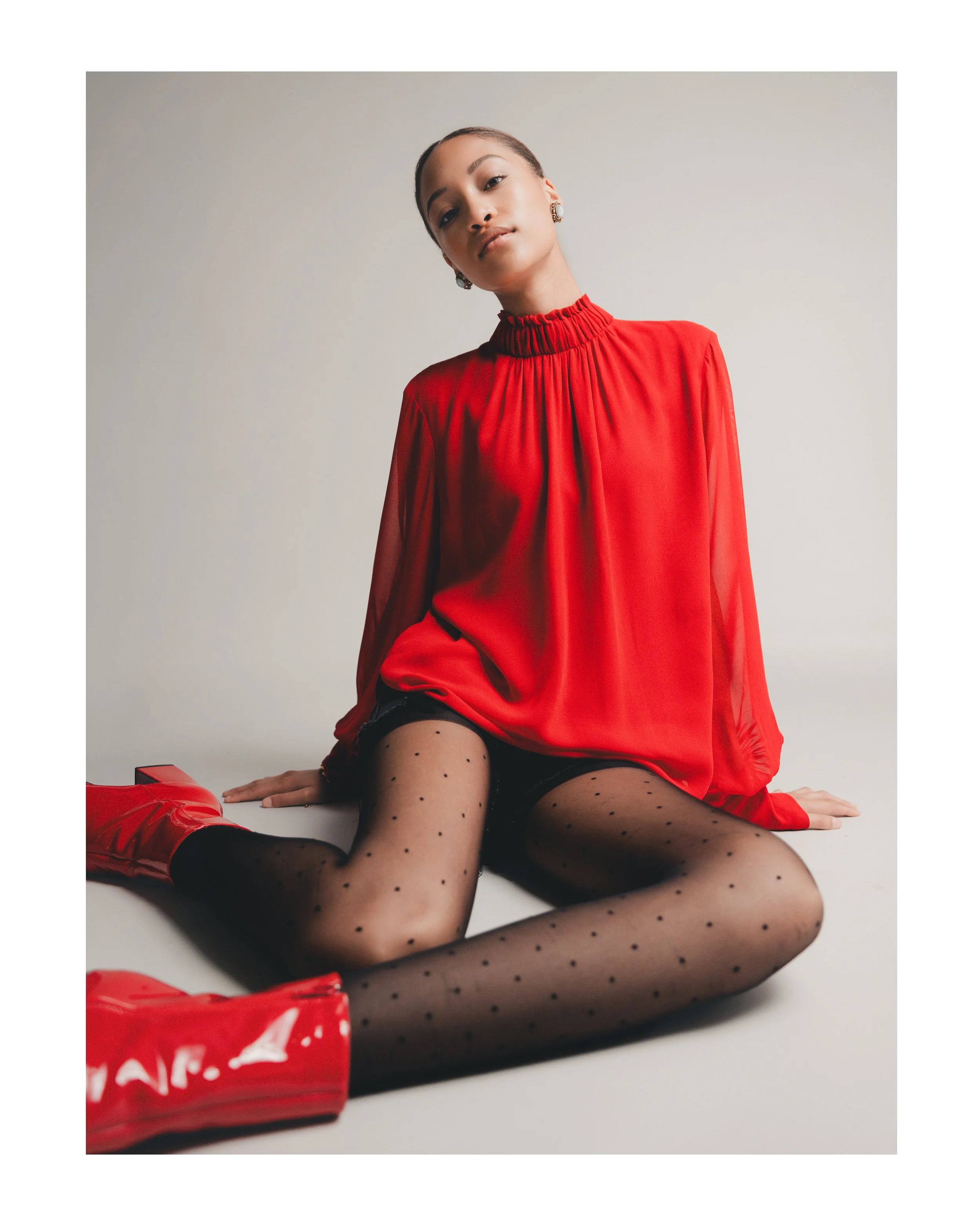 A woman with a confident expression wearing a red blouse, black polka dot stockings, and red shiny over-the-knee boots poses on a plain gray background.